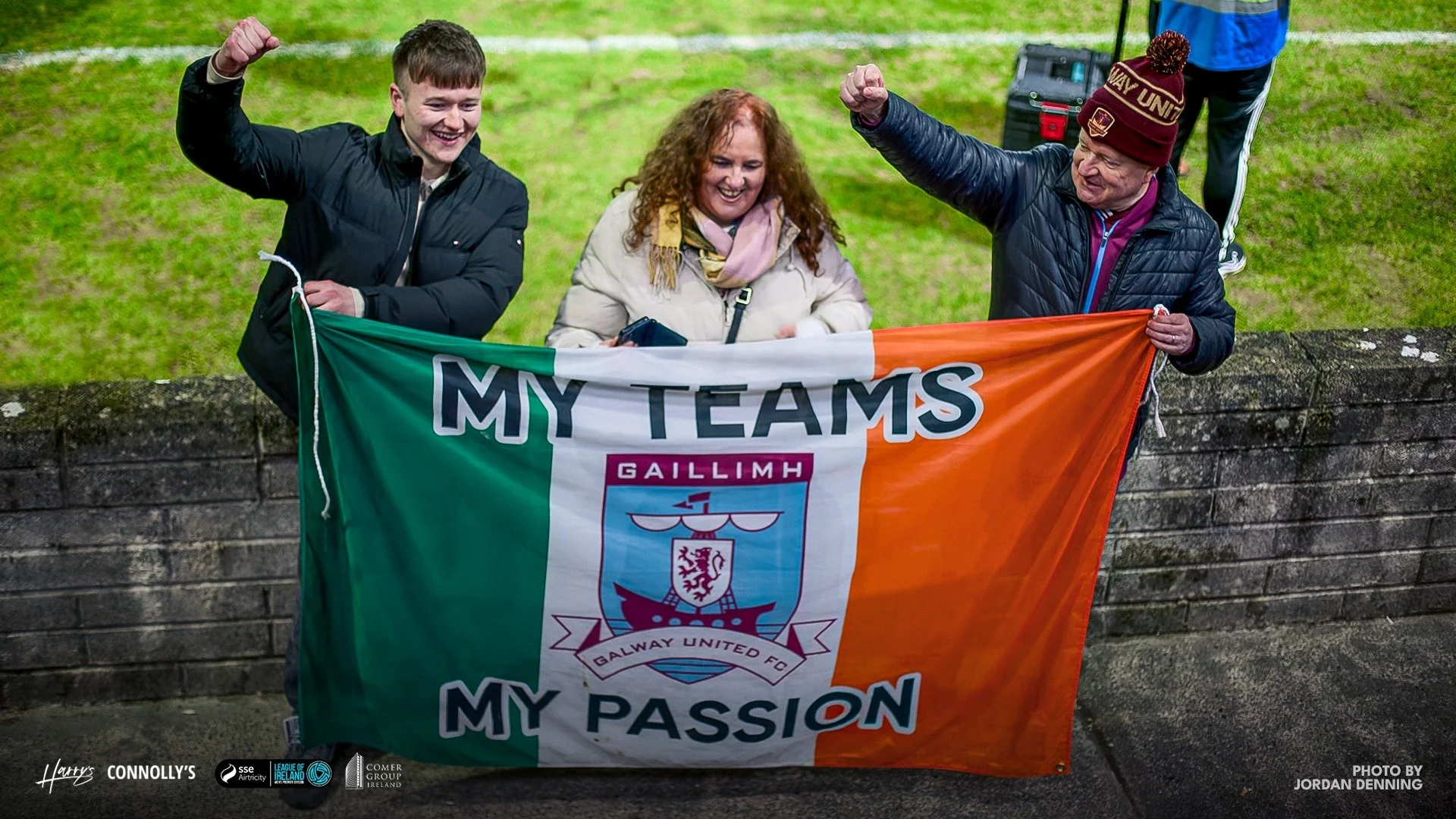 Three people holding an Irish flag with a crest and the words "MY TEAMS, MY PASSION" at a sports event, two of them raising their fists in celebration.