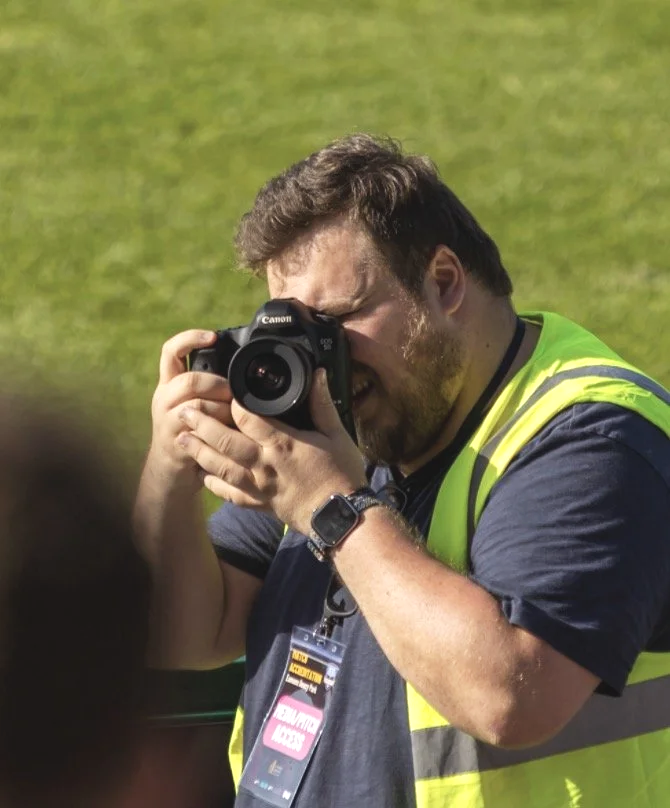 Photographer wearing a yellow safety vest and wristwatch, taking a photo with a Canon camera, outdoors on a sunny day.