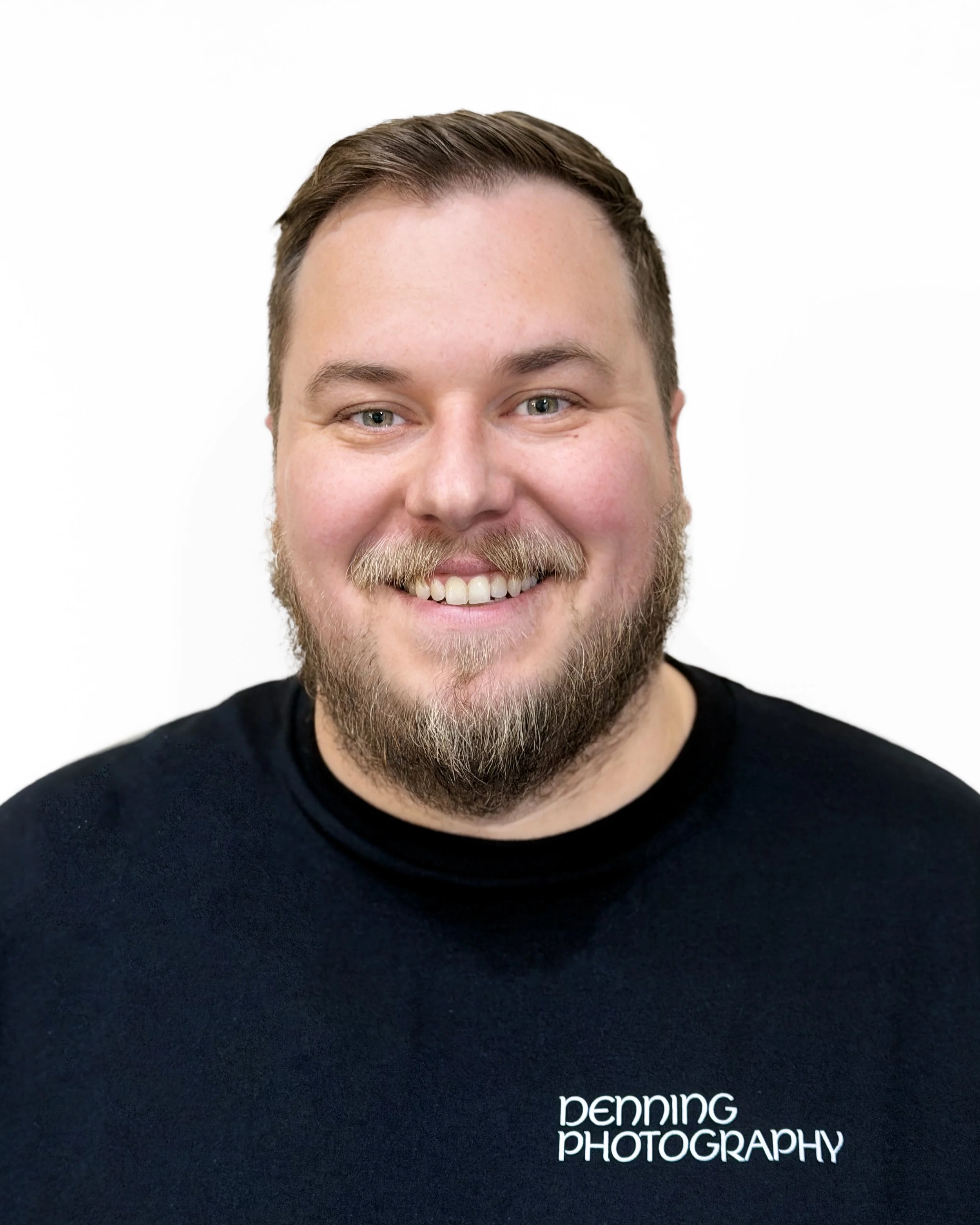 Close-up of a smiling man with short brown hair, blue eyes, and a beard, wearing a black shirt with white text that reads 'denning PHOTOGRAPHY' on a plain white background.