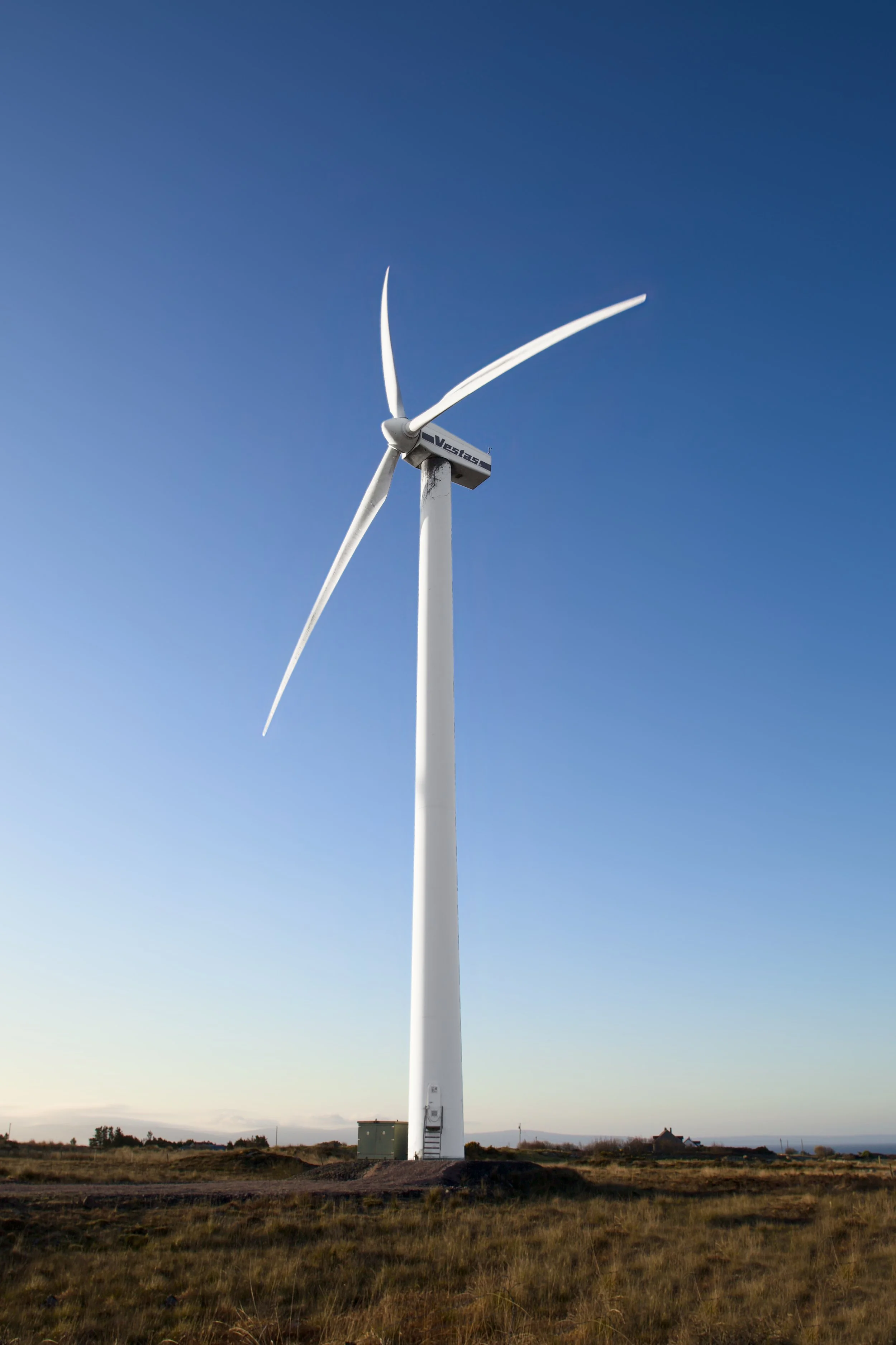 A large white wind turbine stands on open land under a clear blue sky, with distant buildings and trees on the horizon.