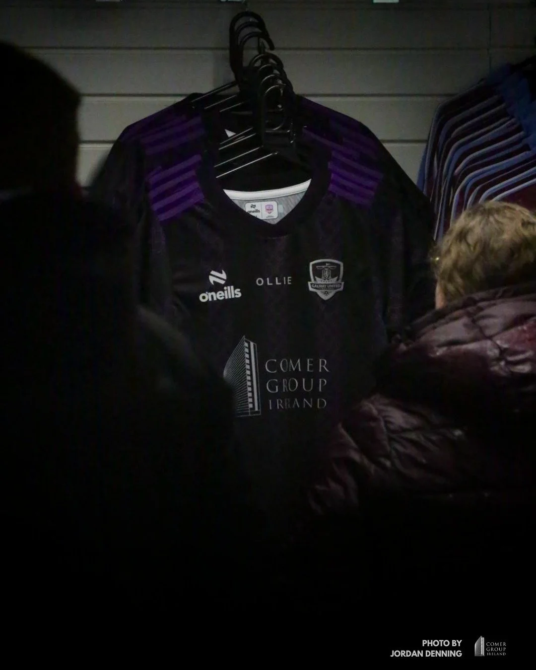 Dark-colored sports jersey hanging on a rack with purple accents, displayed in a retail store, with people observing in front of it.