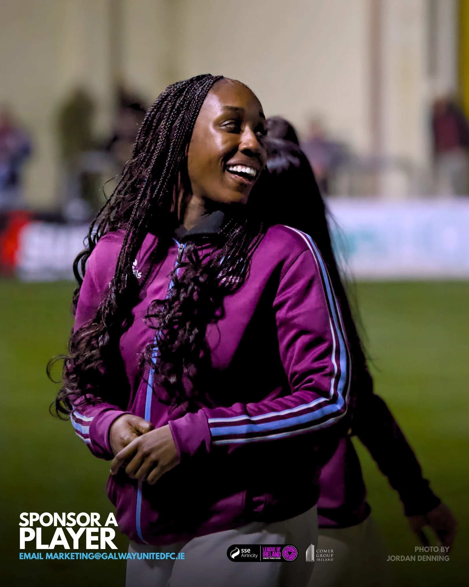A woman with long braided hair smiling at an event, wearing a purple jacket, with a blurred background of people and banners.