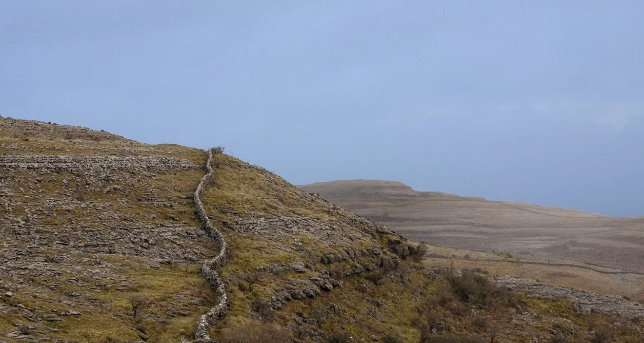 Hilly landscape with rocky slopes and a dry stone wall winding up the hillside under a partly cloudy sky.