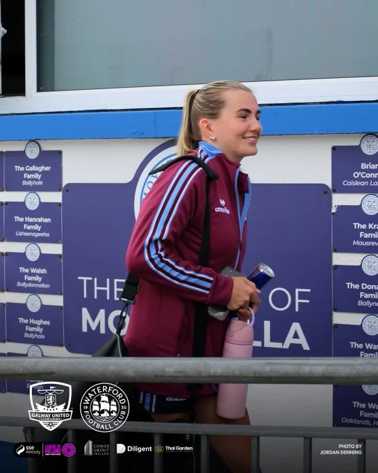 A young woman with blonde hair tied back, wearing a maroon sports jacket with blue and white stripes, holds a pink water bottle and a phone, standing outdoors in front of a signboard with blue and white colors, smiling.