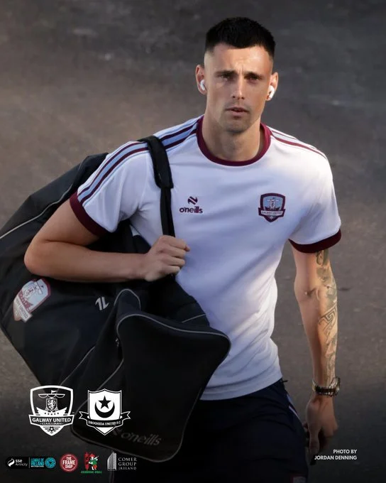 A male athlete carrying a black sports bag over his shoulder, wearing a white sports jersey with maroon trim, and standing on a dirt or gravel surface.