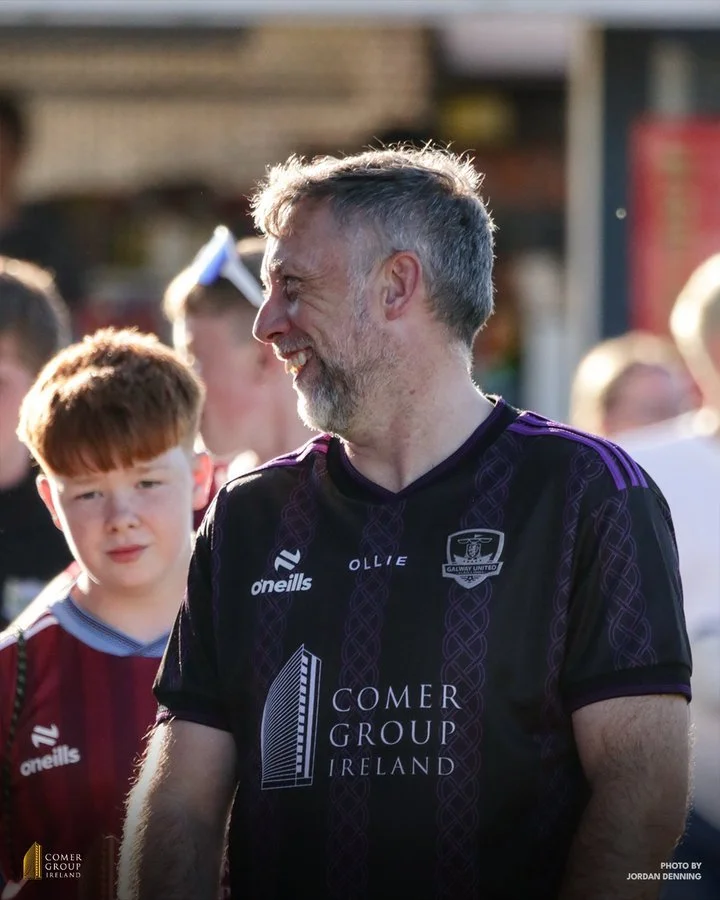 Man with gray beard and short gray hair smiling and looking to the side, wearing a black and purple sports jersey with logos, surrounded by people, outdoors during daytime.