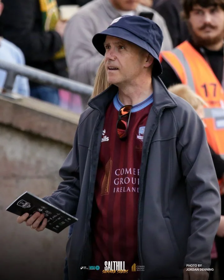 A man wearing a navy bucket hat, maroon sports jersey, and gray jacket holding a booklet at an outdoor sports event, with several spectators behind him.
