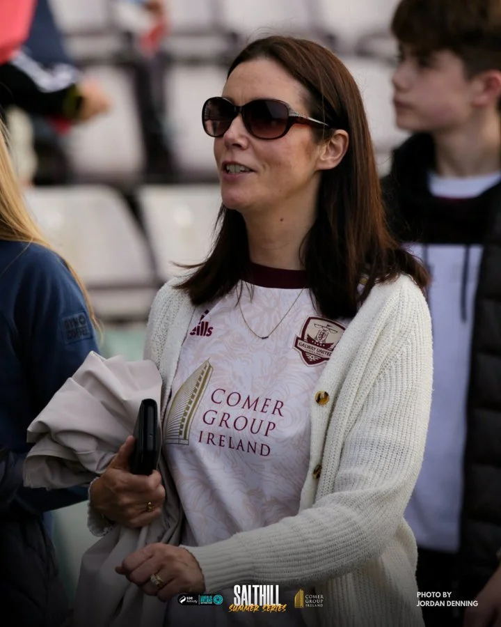 A woman with dark hair and sunglasses wearing a white shirt with 'Comer Group Ireland' logo, holding a beige jacket and a phone, is outdoors at a sporting event, with other people around her.