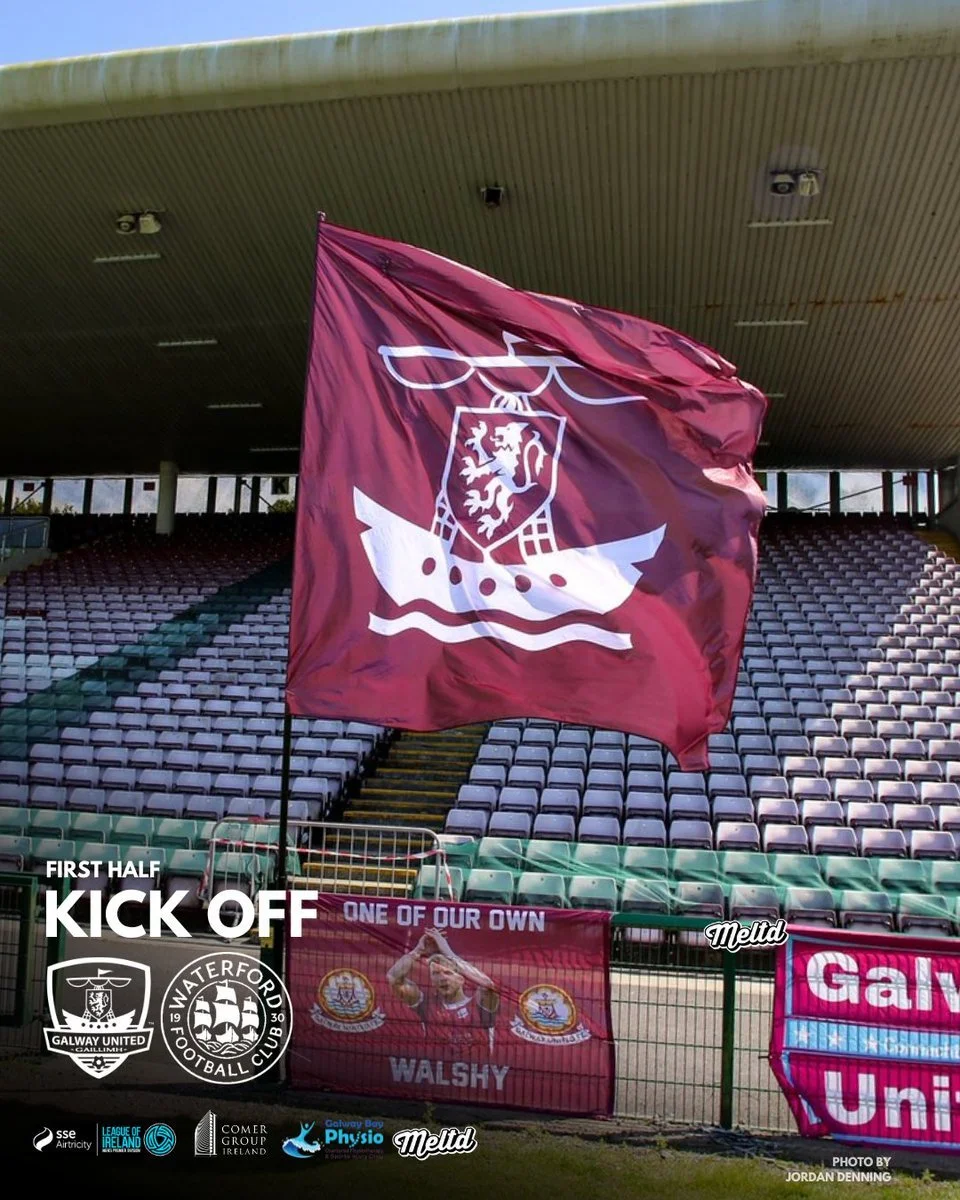 A maroon flag with a white logo of a ship flying a shield with a lion, waving in a stadium with empty seats. In the foreground, there are banners and logos for Galway United and Waterford football clubs, indicating a soccer match kickoff.