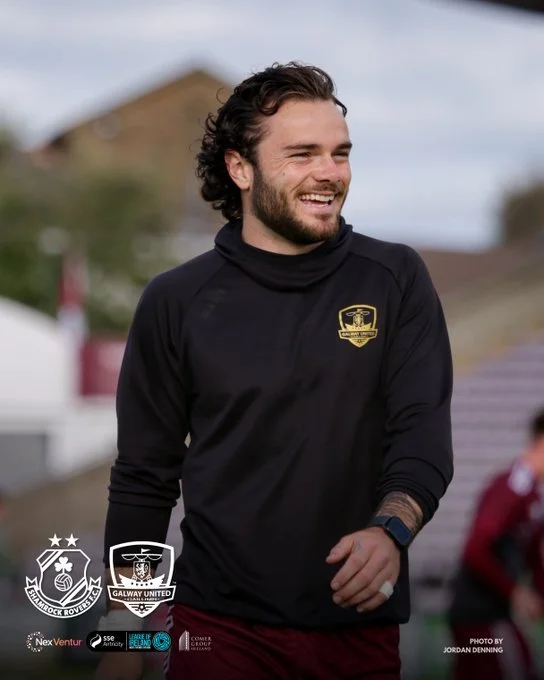 A smiling man with dark curly hair and a beard, wearing a black sports jacket, stands outdoors with a blurred background of buildings and other people.