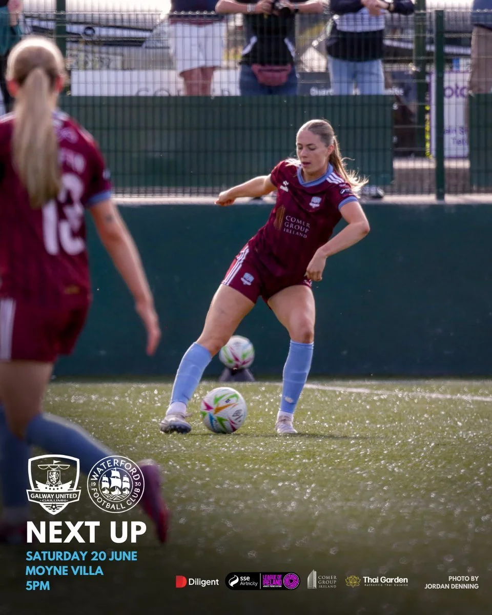 A female soccer player in maroon uniform preparing to kick a soccer ball during a match on a sunny day. Other players and spectators are visible in the background.
