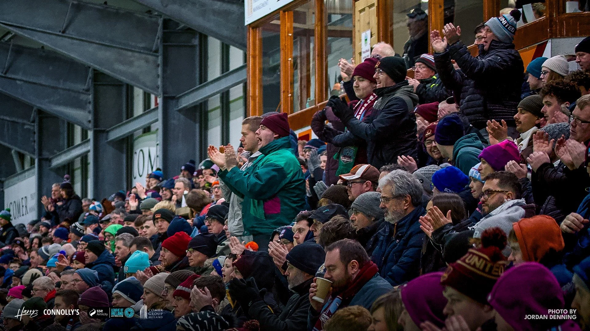 Crowd of sports fans wearing winter clothing at an outdoor event, some standing and clapping, in a stadium with a metal structure and glass windows.