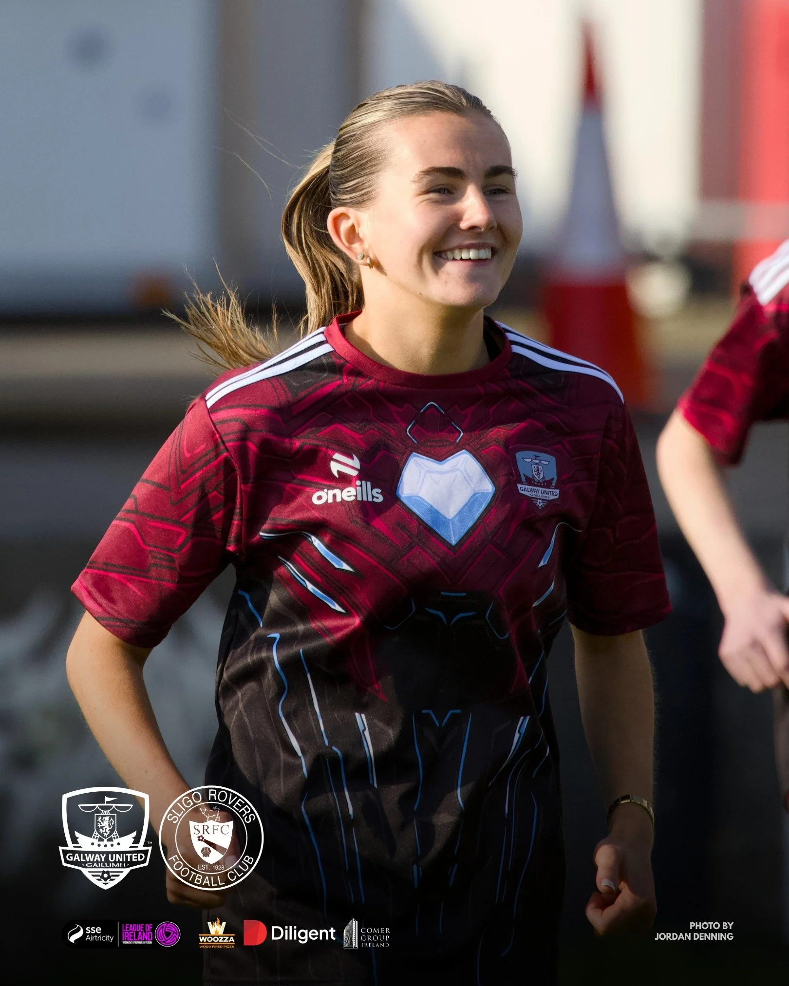 A female soccer player with light skin and blonde hair in a ponytail, smiling and wearing a maroon and black jersey with white and blue accents, standing outdoors during daytime. Logos of Galway United, Sligo Rovers, and various sponsors are visible 