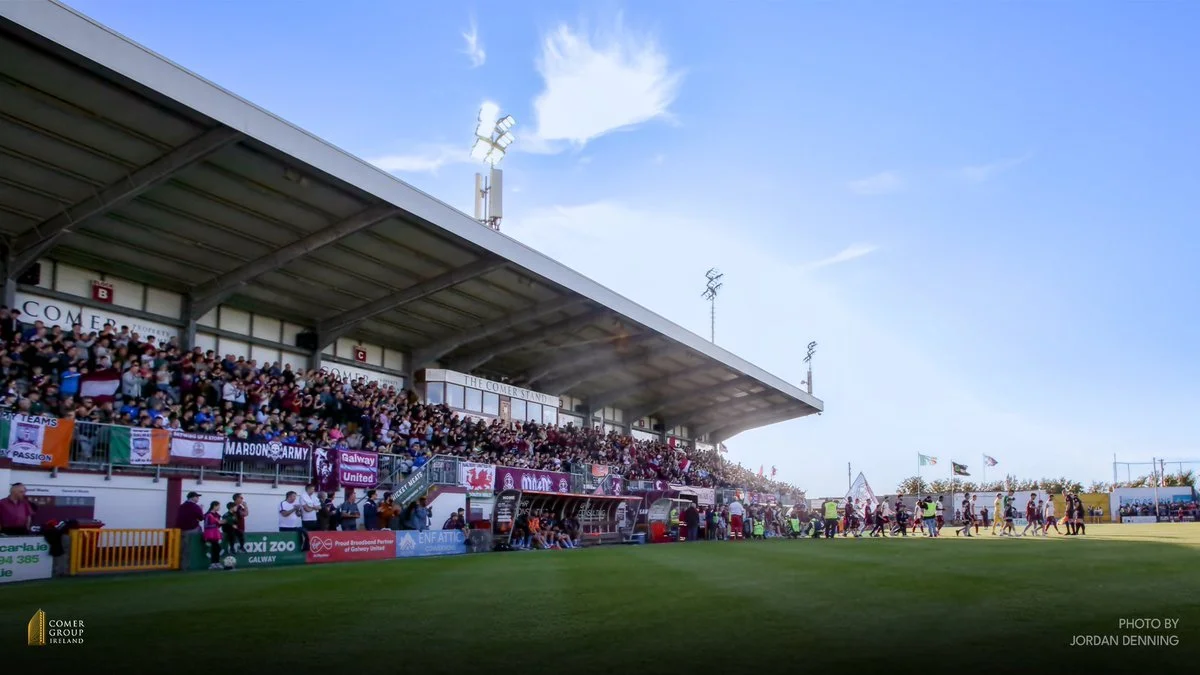 A soccer match at a stadium with a crowd of spectators in the stands, and players on the field. Clear sky with some clouds overhead.