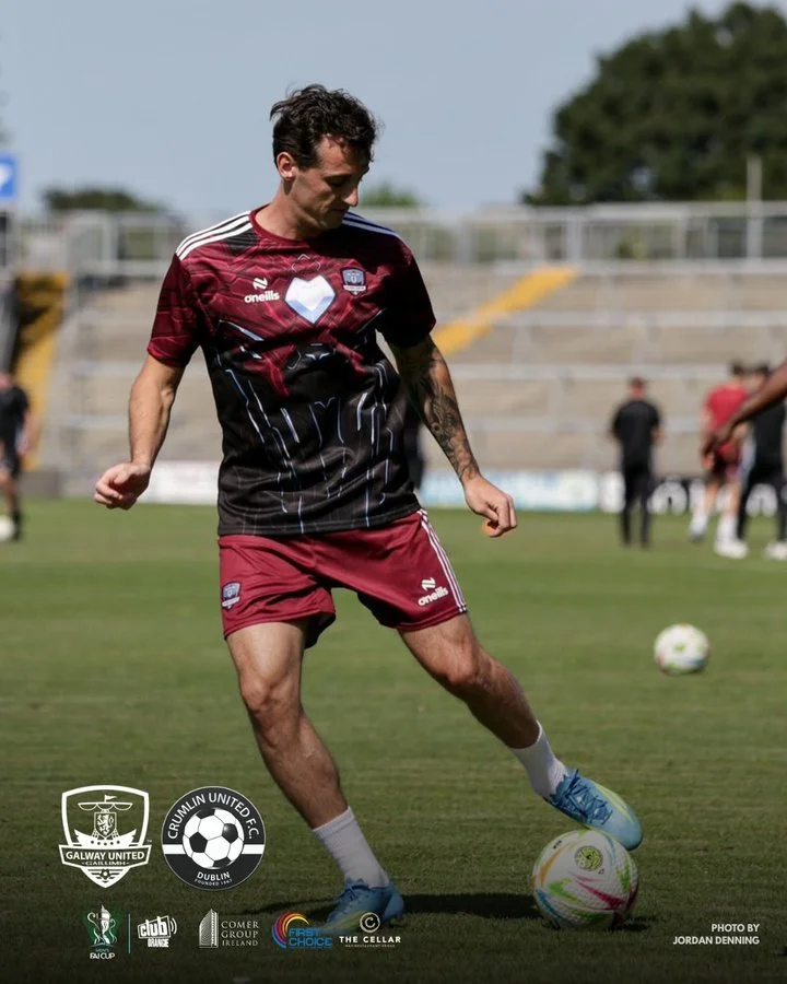 A soccer player practicing on a field, wearing a maroon and black jersey, maroon shorts, and blue cleats, with other people and stadium seats in the background.