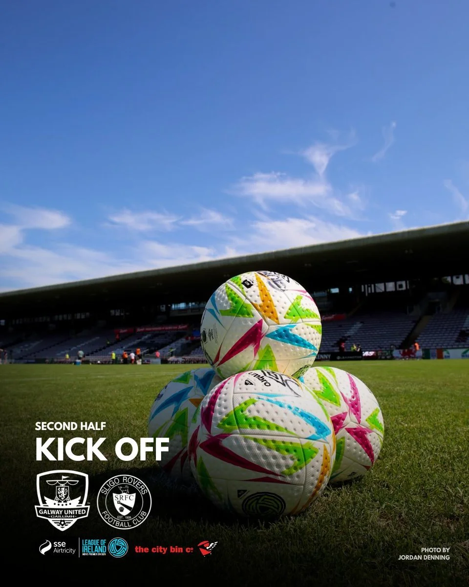 Four colorful soccer balls stacked on a grass field in a stadium with a blue sky and clouds, preparing for the second half kick-off of a match between Galway United and Sligo Rovers.