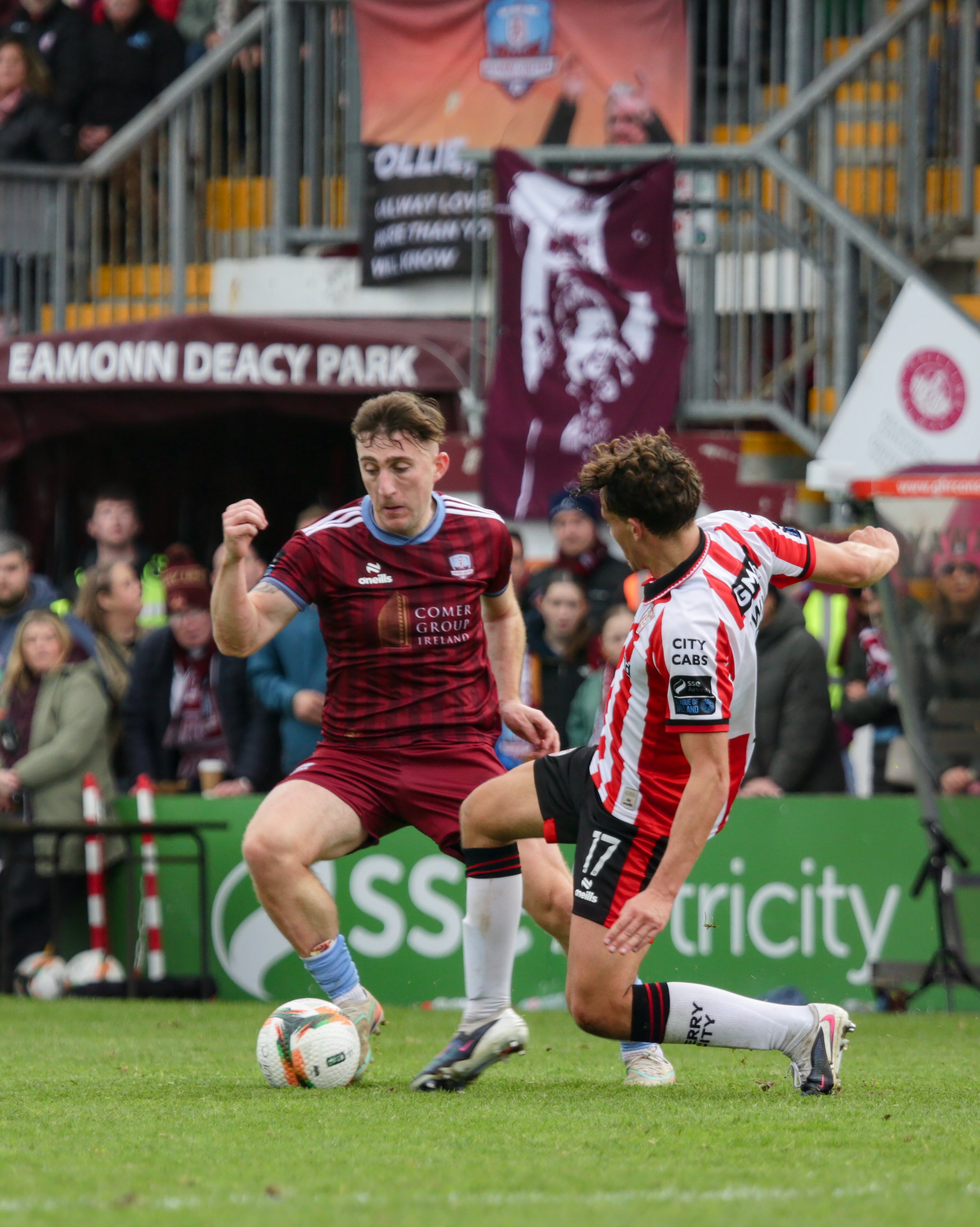 Two soccer players compete for the ball during a match at Eamonn Deacy Park. One player in a maroon jersey and the other in a white and red striped jersey. There are spectators in the background and banners visible around the field.