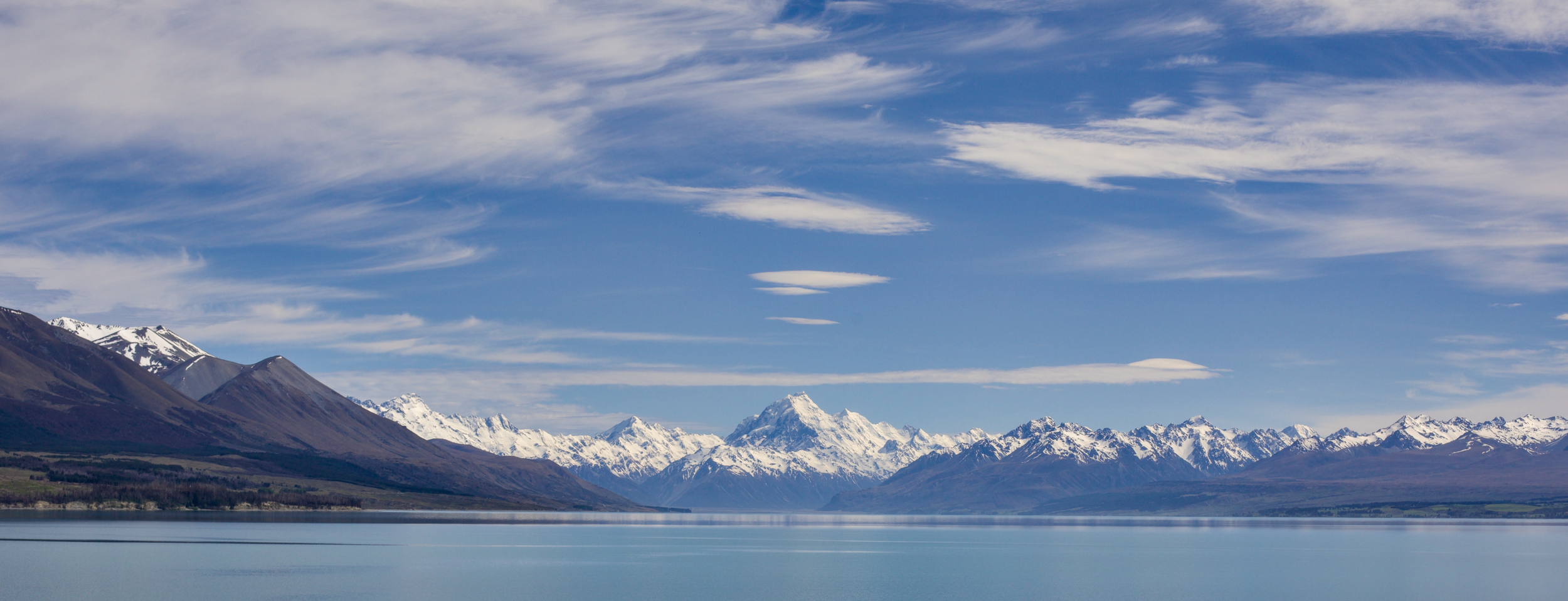 A serene landscape of snow-capped mountains beside a calm lake under a blue sky with white clouds.