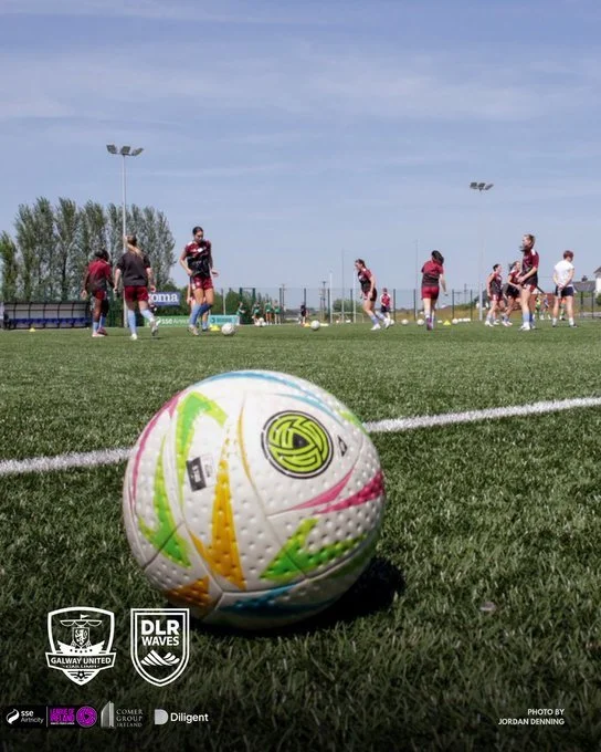 Close-up of a colorful soccer ball on a grassy field with a group of players practicing in the background on a sunny day.