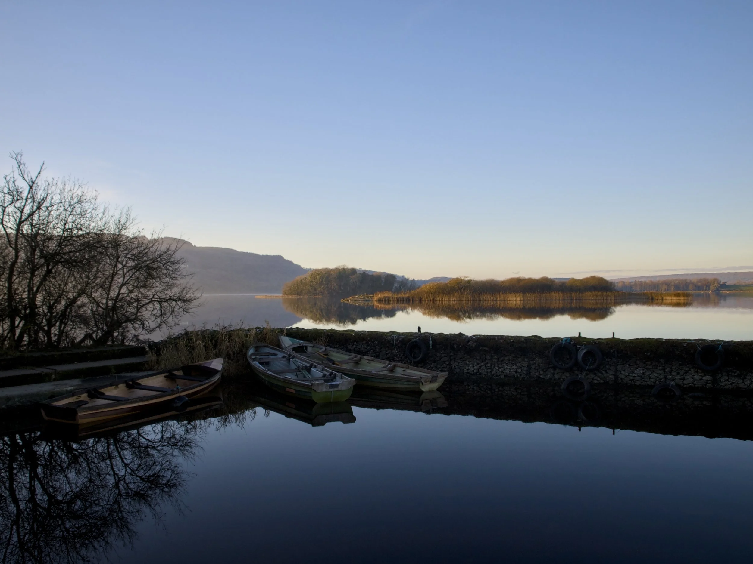 Boats resting along the shoreline of a calm river or lake, with trees and hills in the distance, under a clear blue sky.