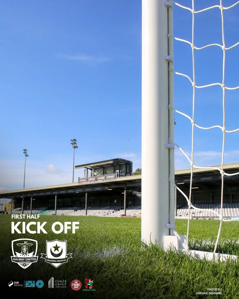 Soccer field with goalpost in the foreground and empty stadium stands in the background under a blue sky, advertising first-half kick-off for a match between Galway United and Drogheda United.