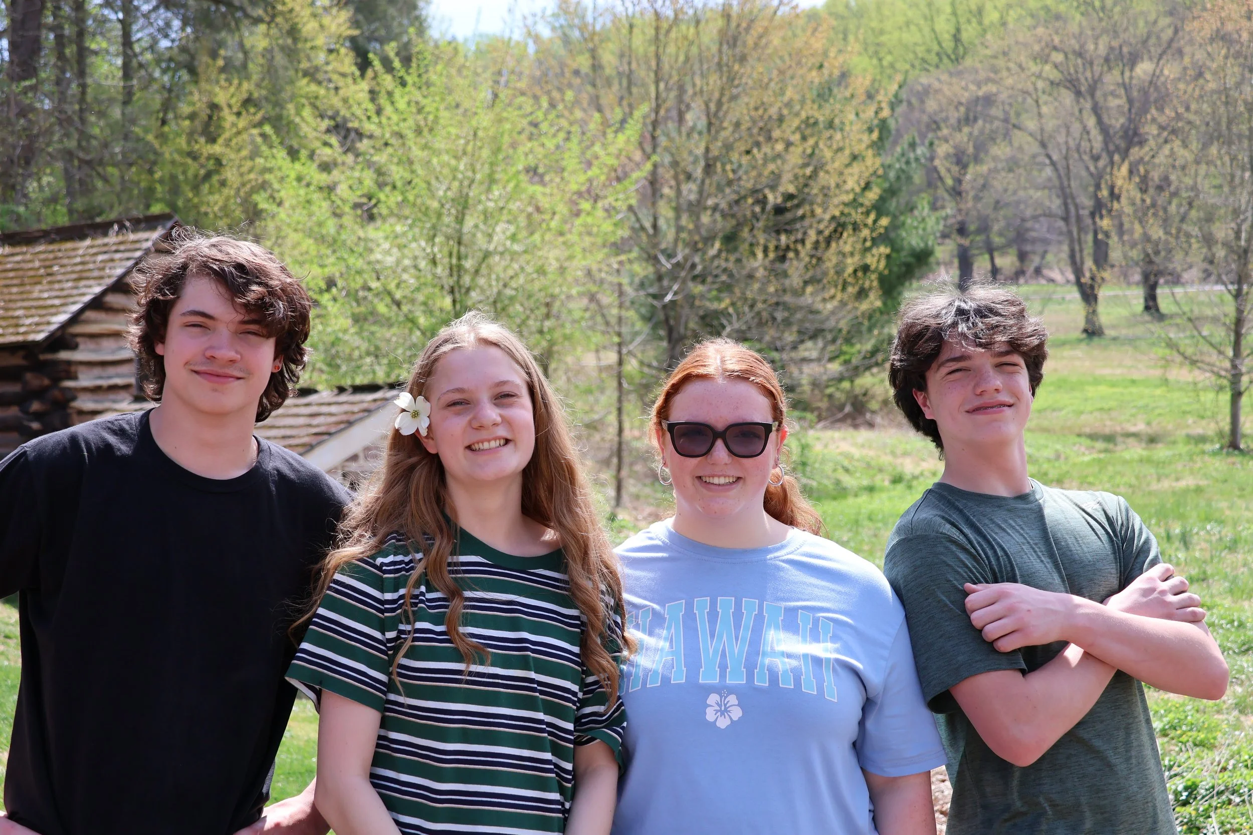 Group of four smiling teenagers outdoors on a sunny day with trees and a rustic building in the background.