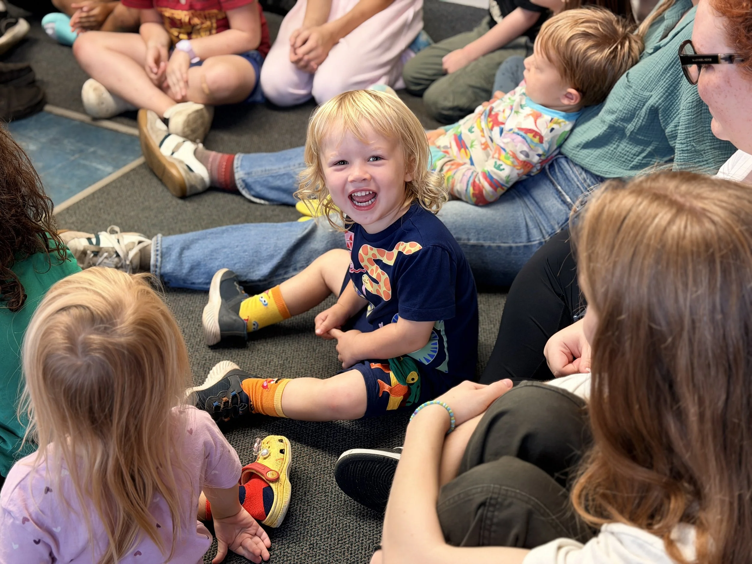 A group of children and adults sitting on the floor in a circle. The focus is on a smiling, blonde boy wearing a Mickey Mouse shirt and colorful socks. The children are engaged in an activity, and the setting appears to be a classroom or playroom.