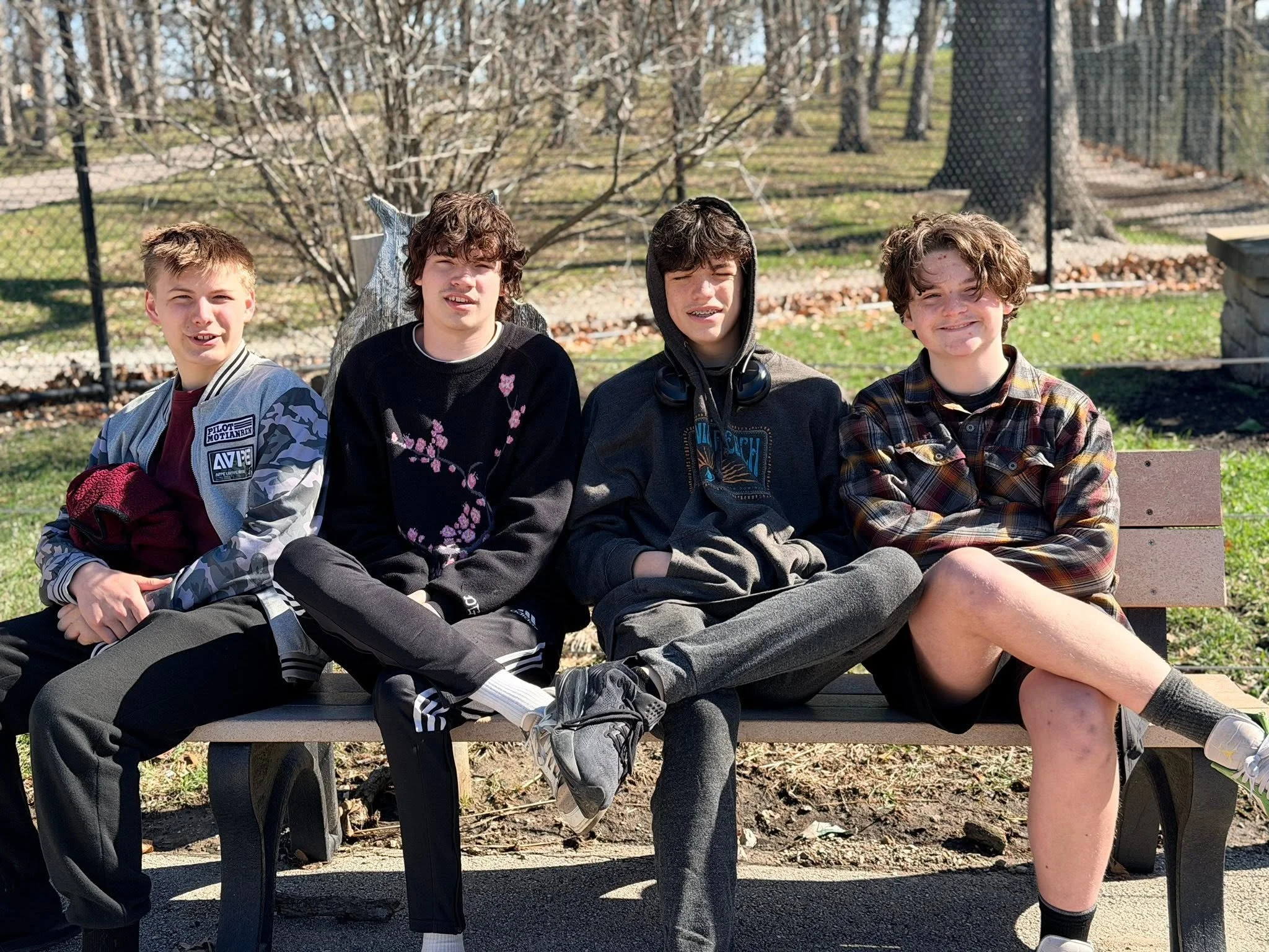Four boys sitting on a park bench outdoors on a sunny day, with trees and a fenced area in the background.