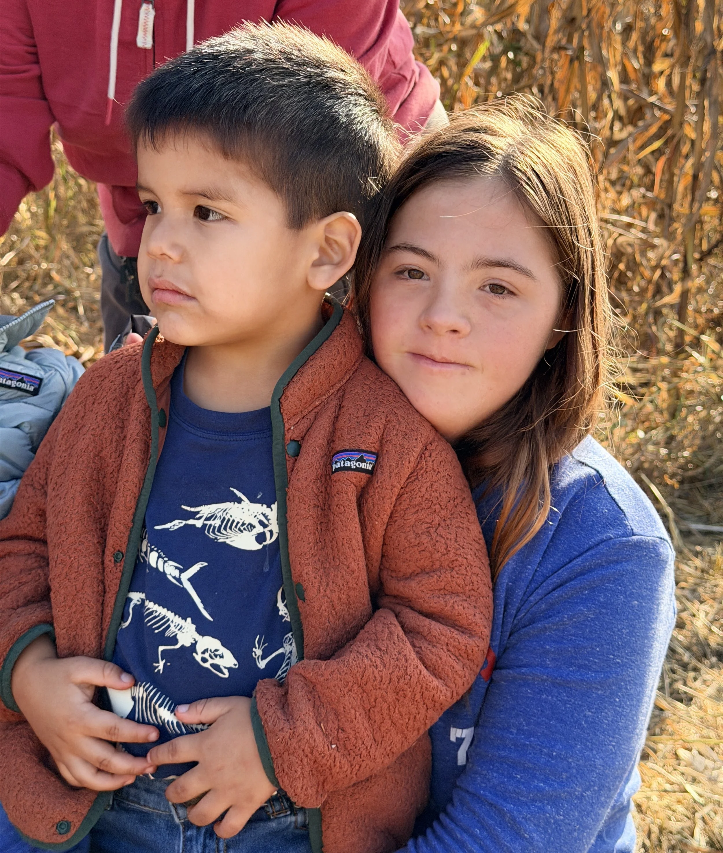 A young boy and girl in an outdoor field during fall, with the girl hugging the boy from behind. The boy has short dark hair and is wearing a Patagonia jacket over a navy shirt with a white dinosaur skeleton graphic. The girl has long brown hair, light skin, and is wearing a blue jacket. The background shows dried plants and golden foliage.