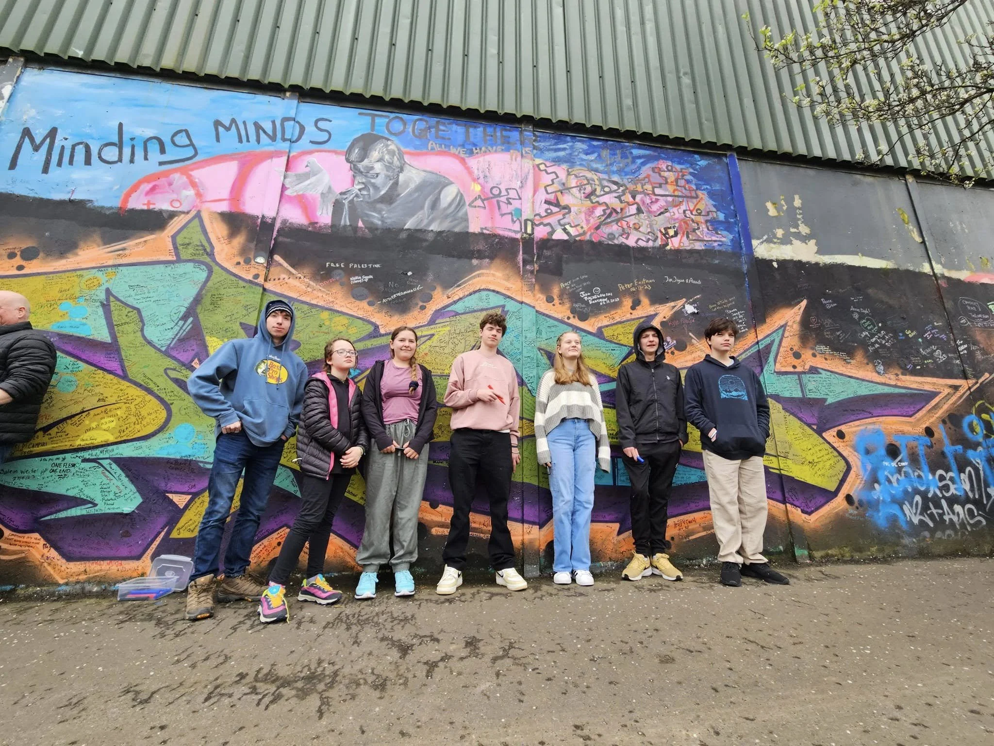 A group of young people standing in front of a large colorful mural on a wall. The mural depicts a person with glasses and a hoodie, surrounded by abstract and graffiti-style elements. The phrase "MINDING MINDS TOGETHER" is written at the top of the mural.