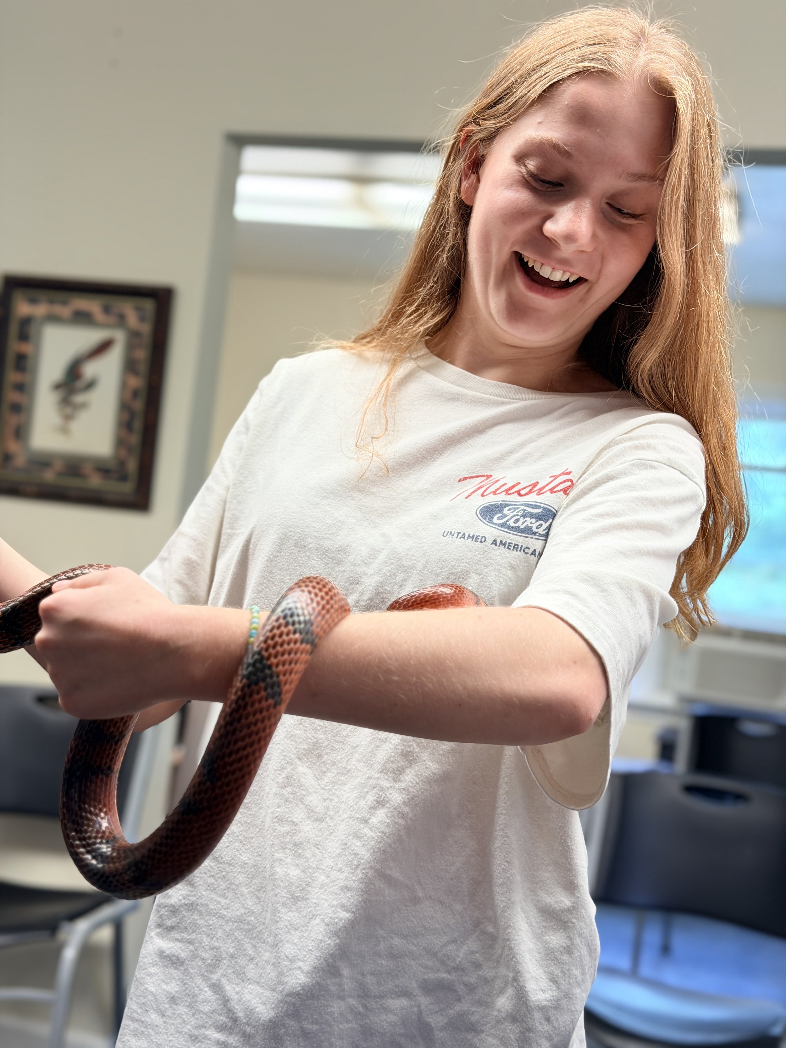 A young woman with long red hair wearing a white t-shirt with a Ford logo, holding a snake in a room with beige walls and framed pictures, smiling and looking down at the snake.