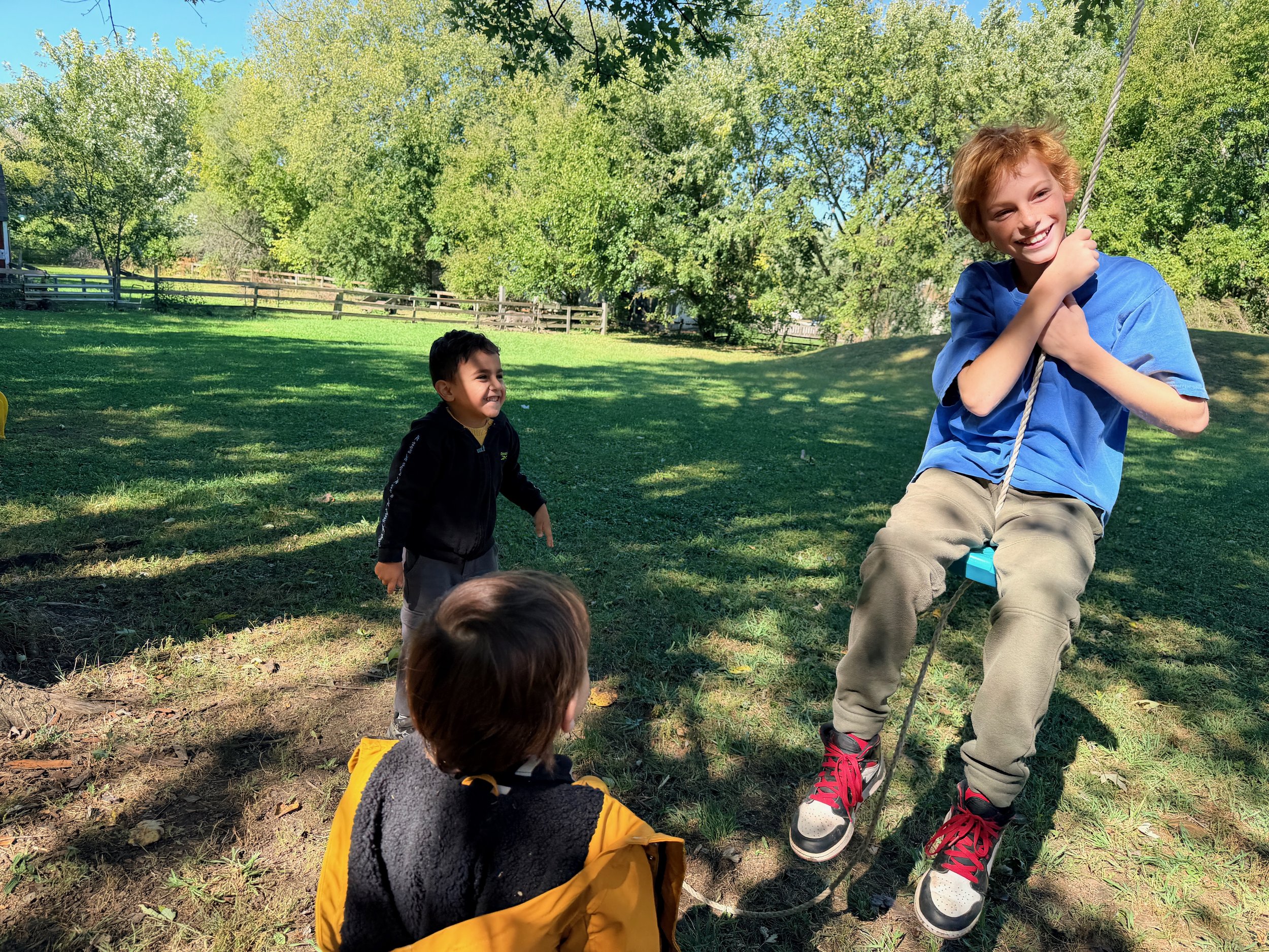 Three children playing outdoors on a sunny day, with green trees in the background. One boy with red hair is smiling and sitting on a swing, holding the ropes. Two other children are sitting on the ground watching him.