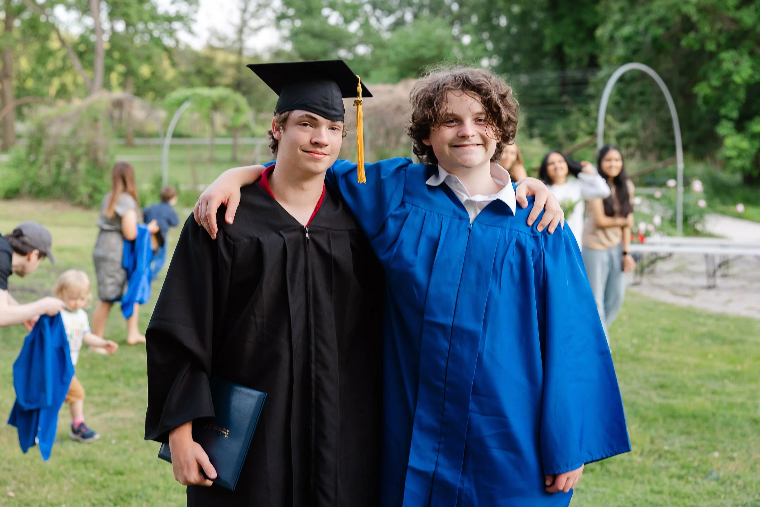 Two young men in graduation gowns and caps with a wooded outdoor setting in the background. They have their arms around each other, smiling.