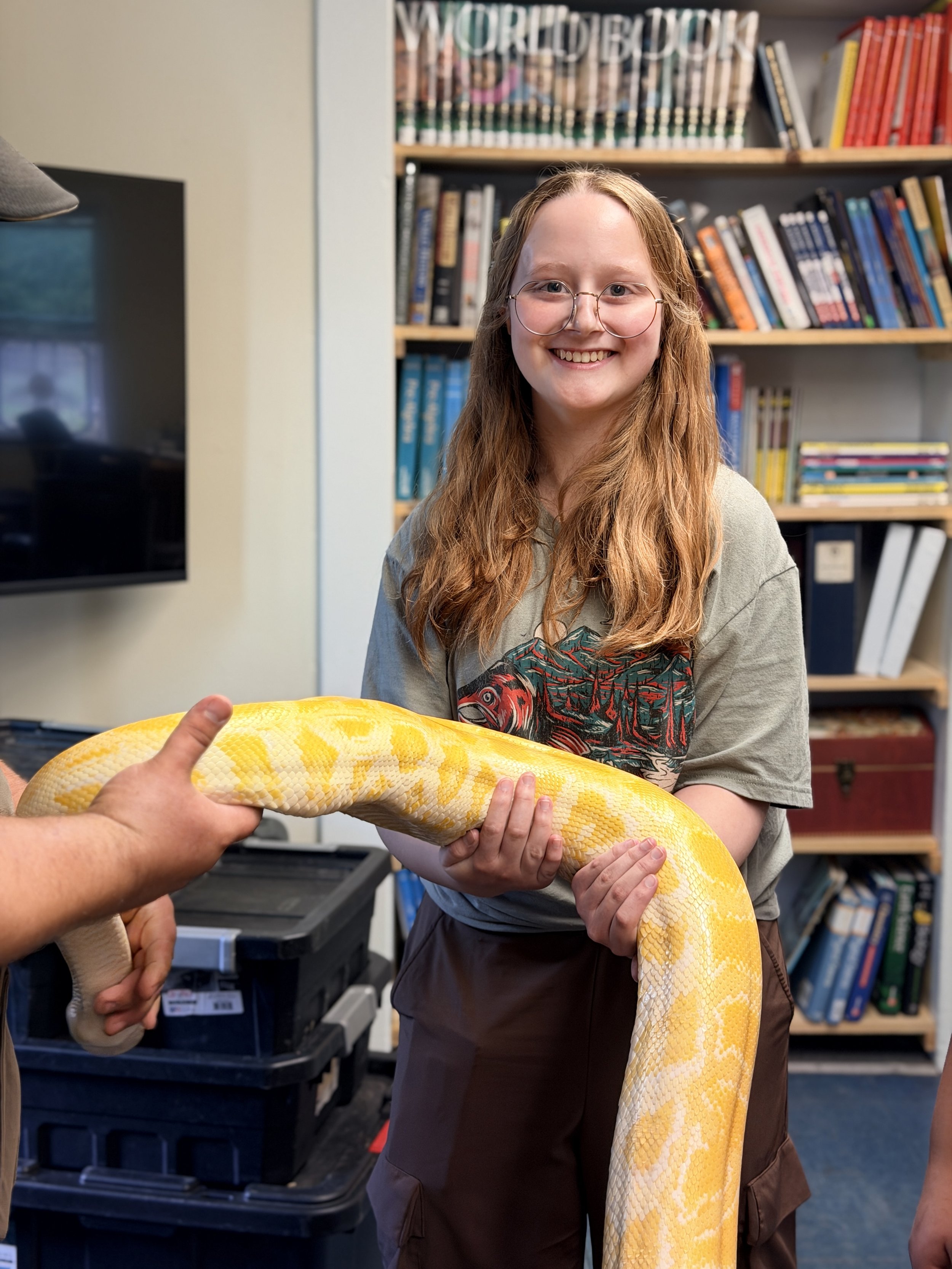 A young woman with long red hair, glasses, and a graphic t-shirt, smiling while holding a large yellow and white snake. The snake is being handed over to her by a person whose hand and part of arm are visible. In the background, there are bookshelves