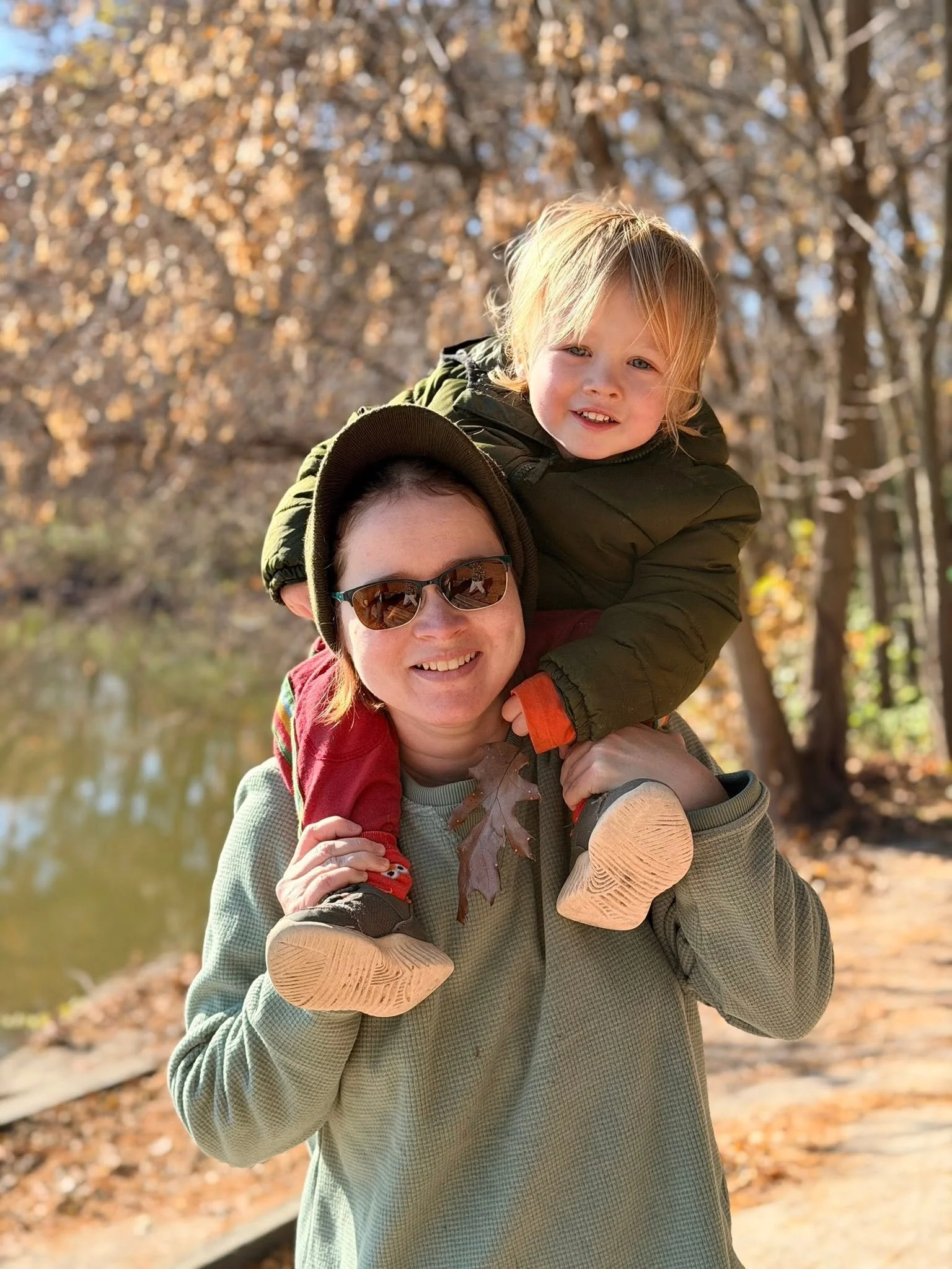 A woman carrying a young child on her shoulders outside during autumn, with trees and fallen leaves in the background.