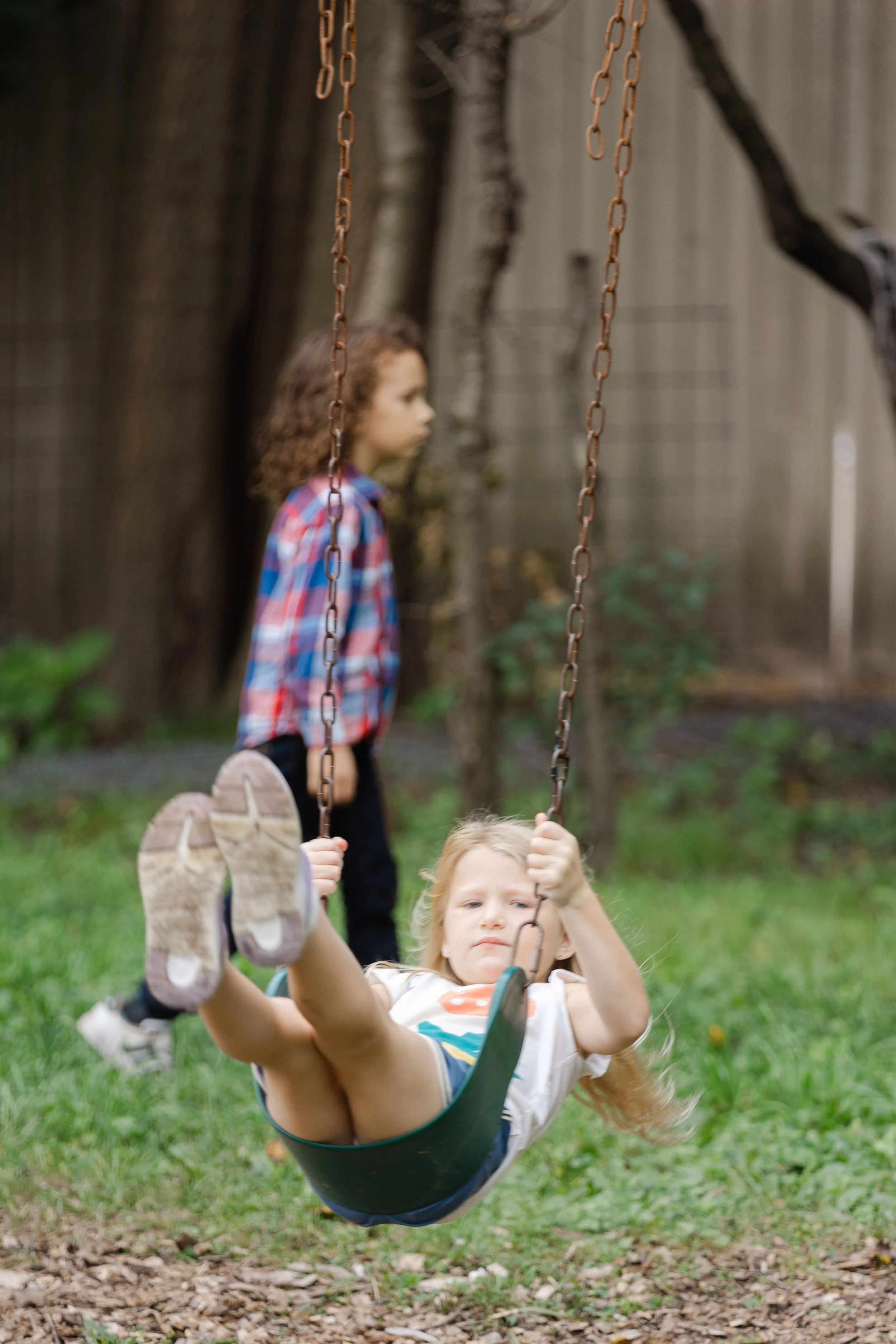Two young children playing on a swing set outdoors. One child is sitting on the swing and smiling, while the other is standing nearby. The background features trees and a wooden fence.