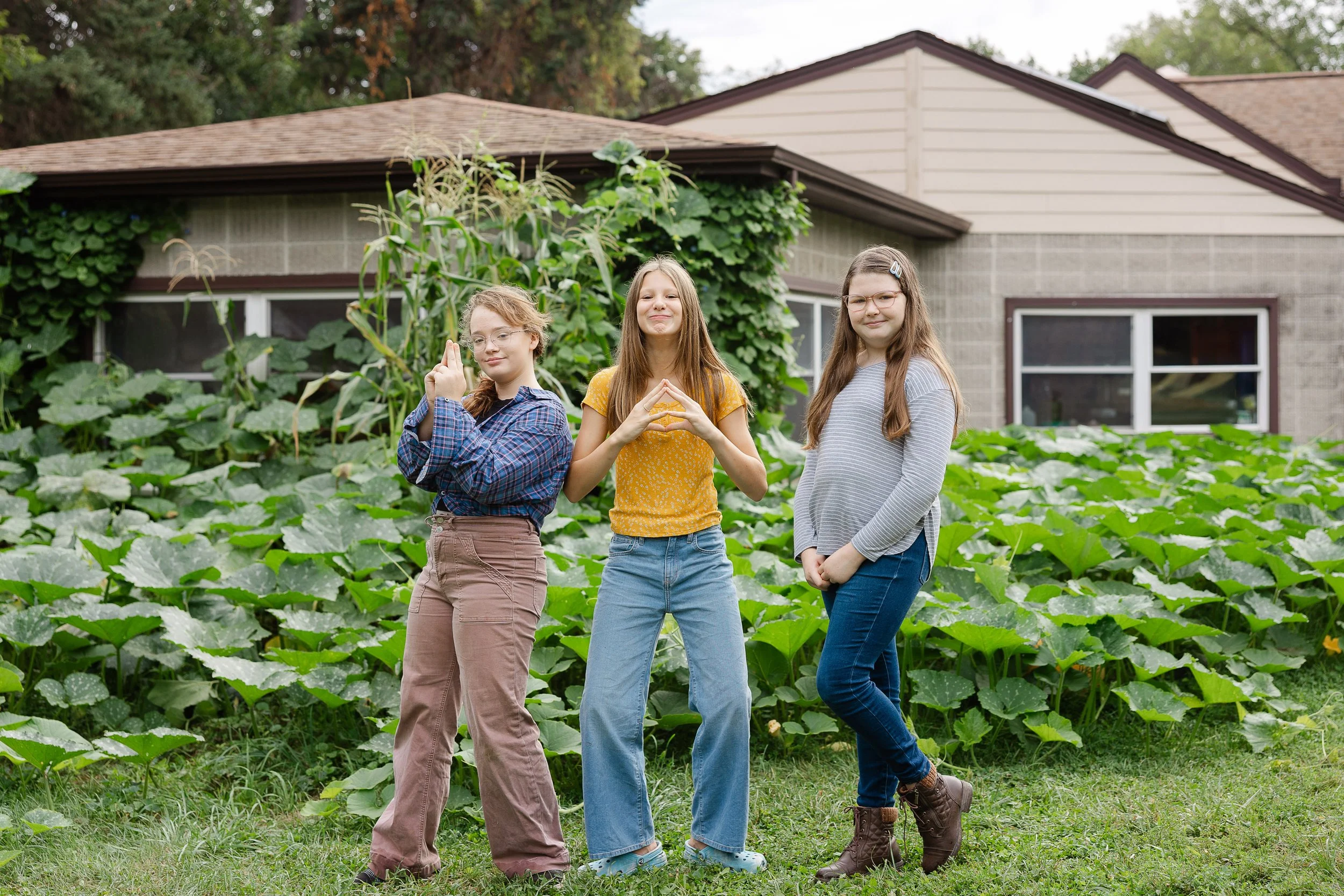 Three young girls stand outdoors in front of a lush garden filled with large green leaves, with a house in the background. They are posing with confident expressions and hand gestures.