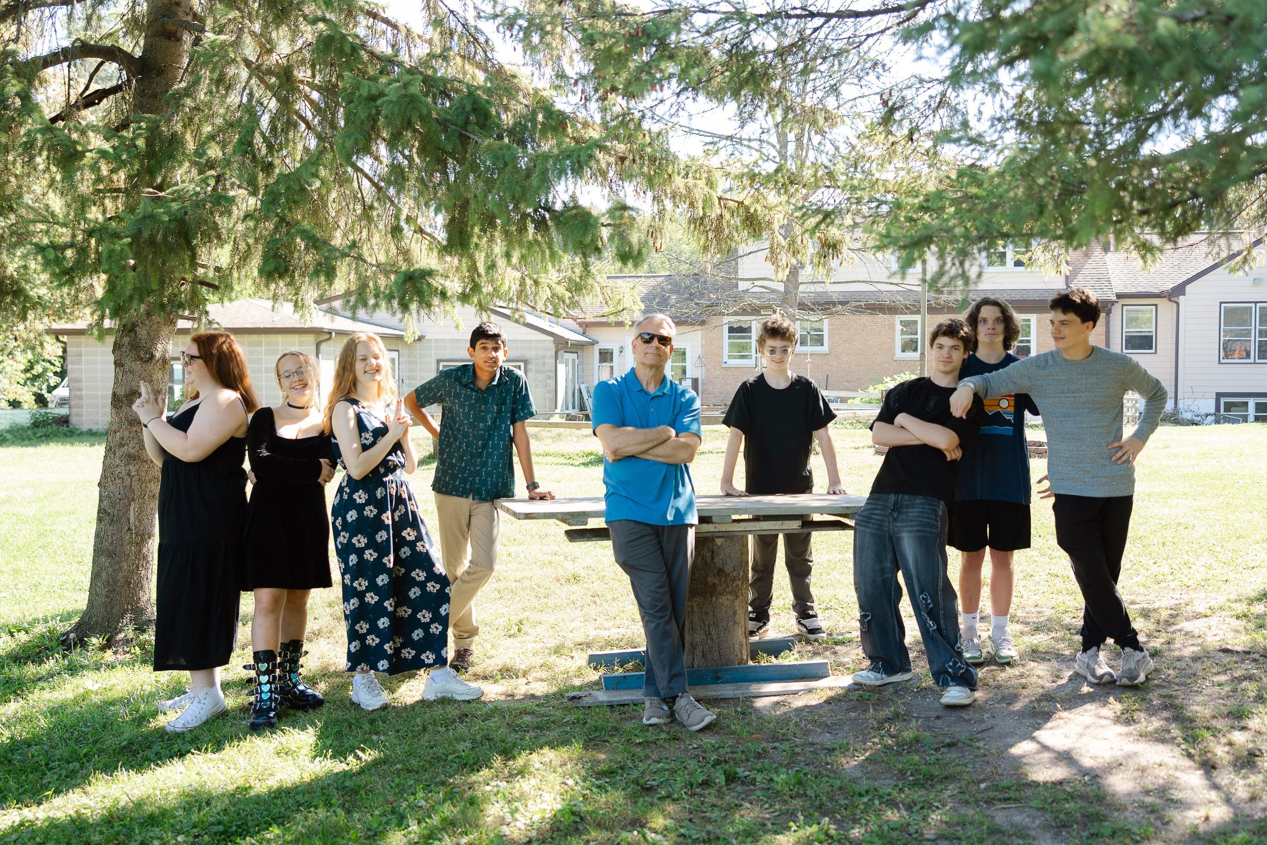 A diverse group of nine young people and one adult man stand outdoors near a large tree on a sunny day, with suburban houses in the background, gathered around a picnic table.
