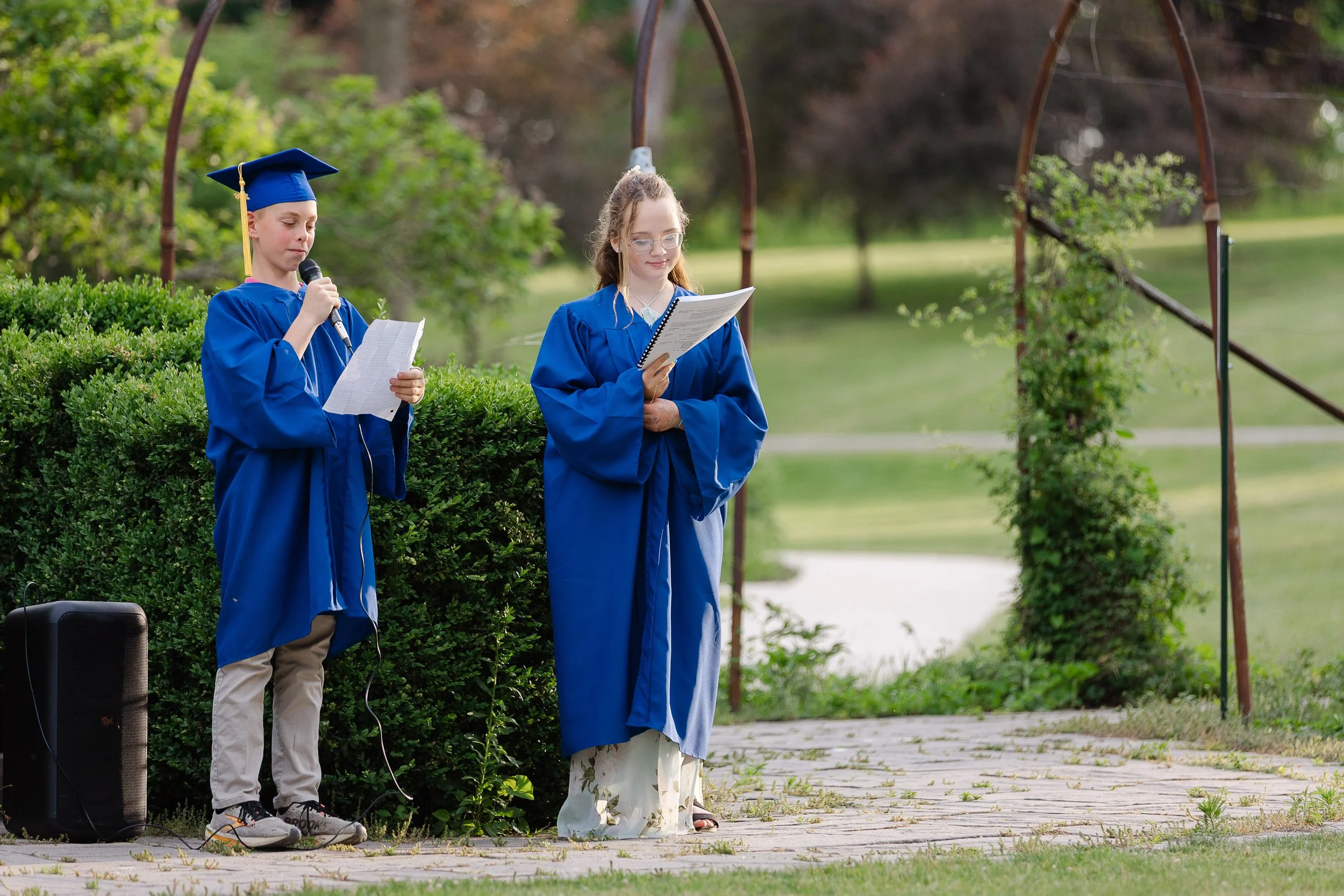 A boy and girl in blue caps and gowns standing outdoors, with the boy speaking into a microphone and the girl holding a paper and reading, during a graduation ceremony.
