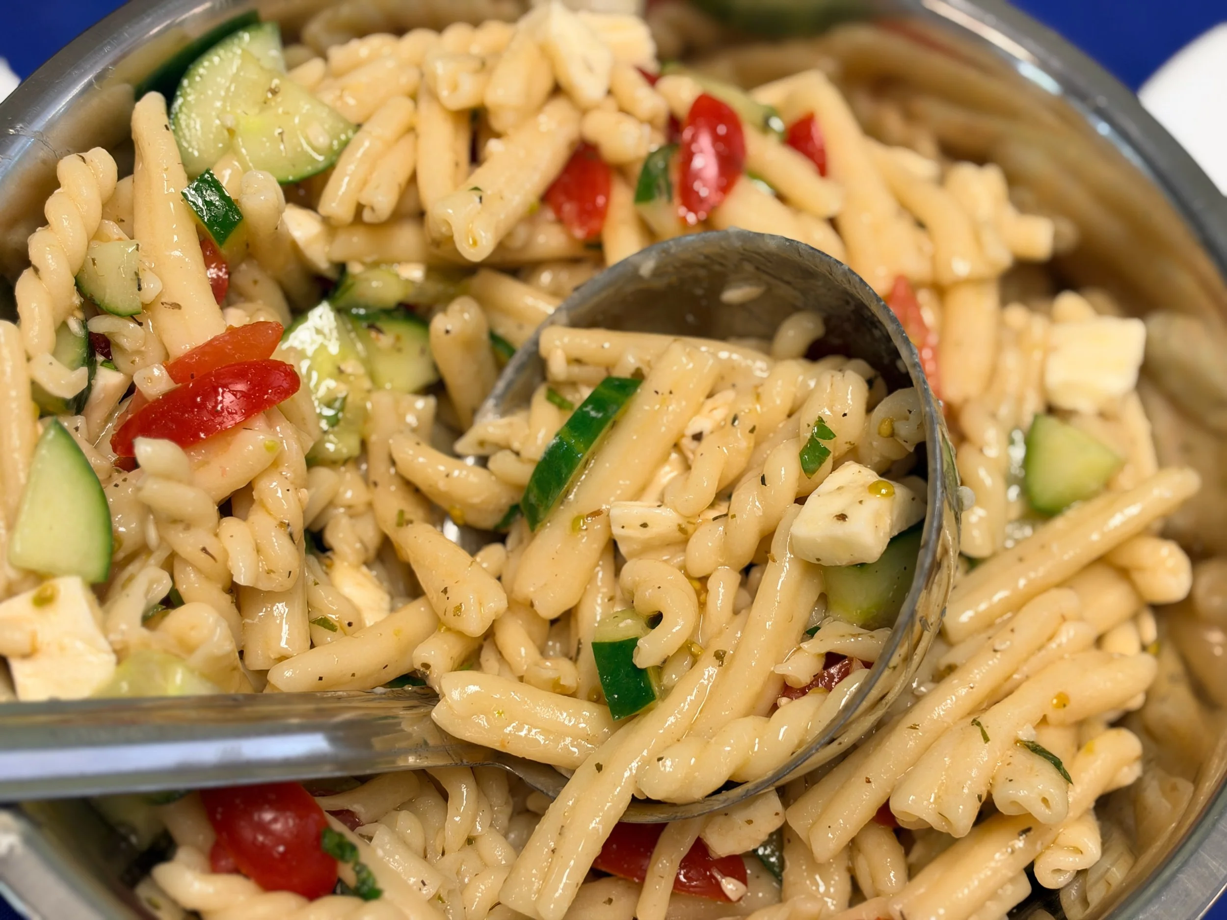 A close-up of a pasta salad with rotini pasta, cherry tomatoes, cucumber slices, chopped green onions, and herbs in a clear glass dish.