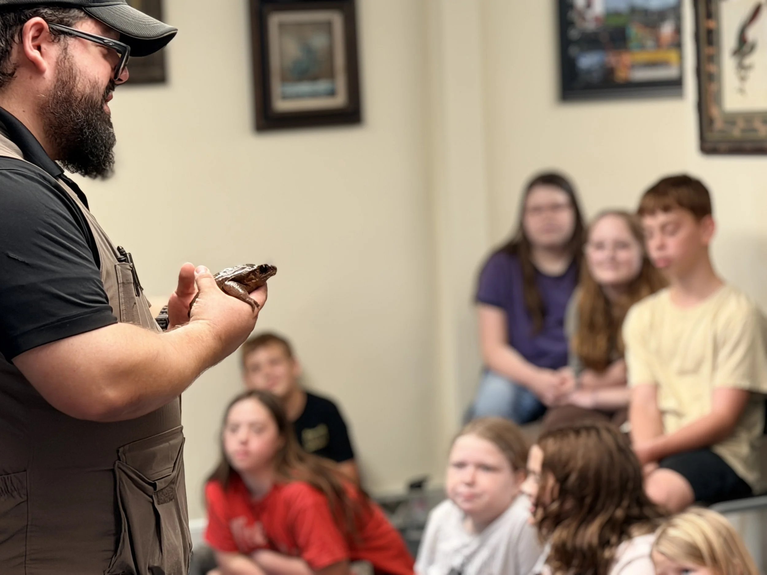 Man holding a small animal or reptile, possibly a gecko, in front of a group of children seated and watching attentively.