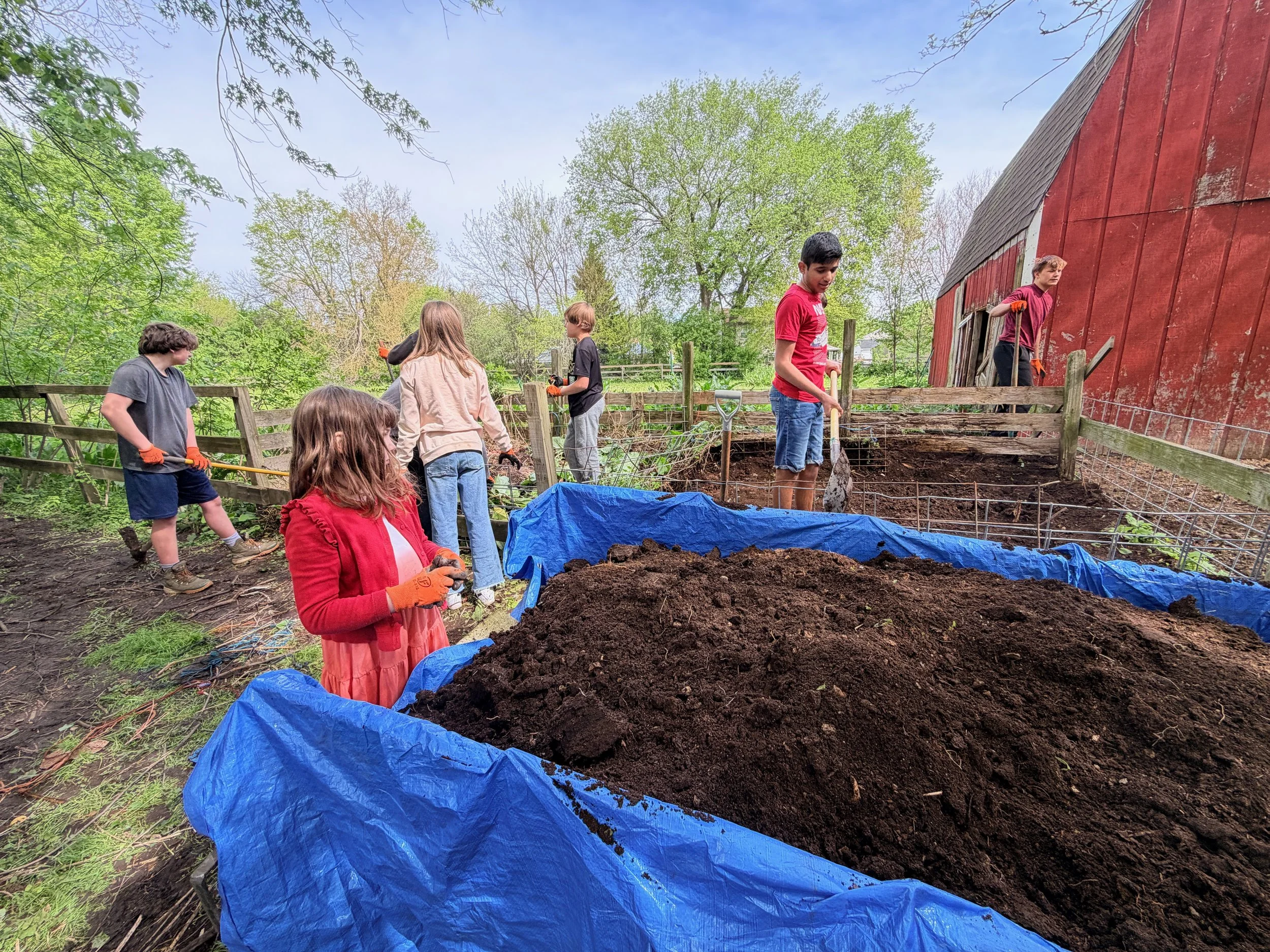 Children and teenagers working together outdoors in a garden or yard; some are holding gardening tools near a large pile of soil, with a red barn and trees in the background.