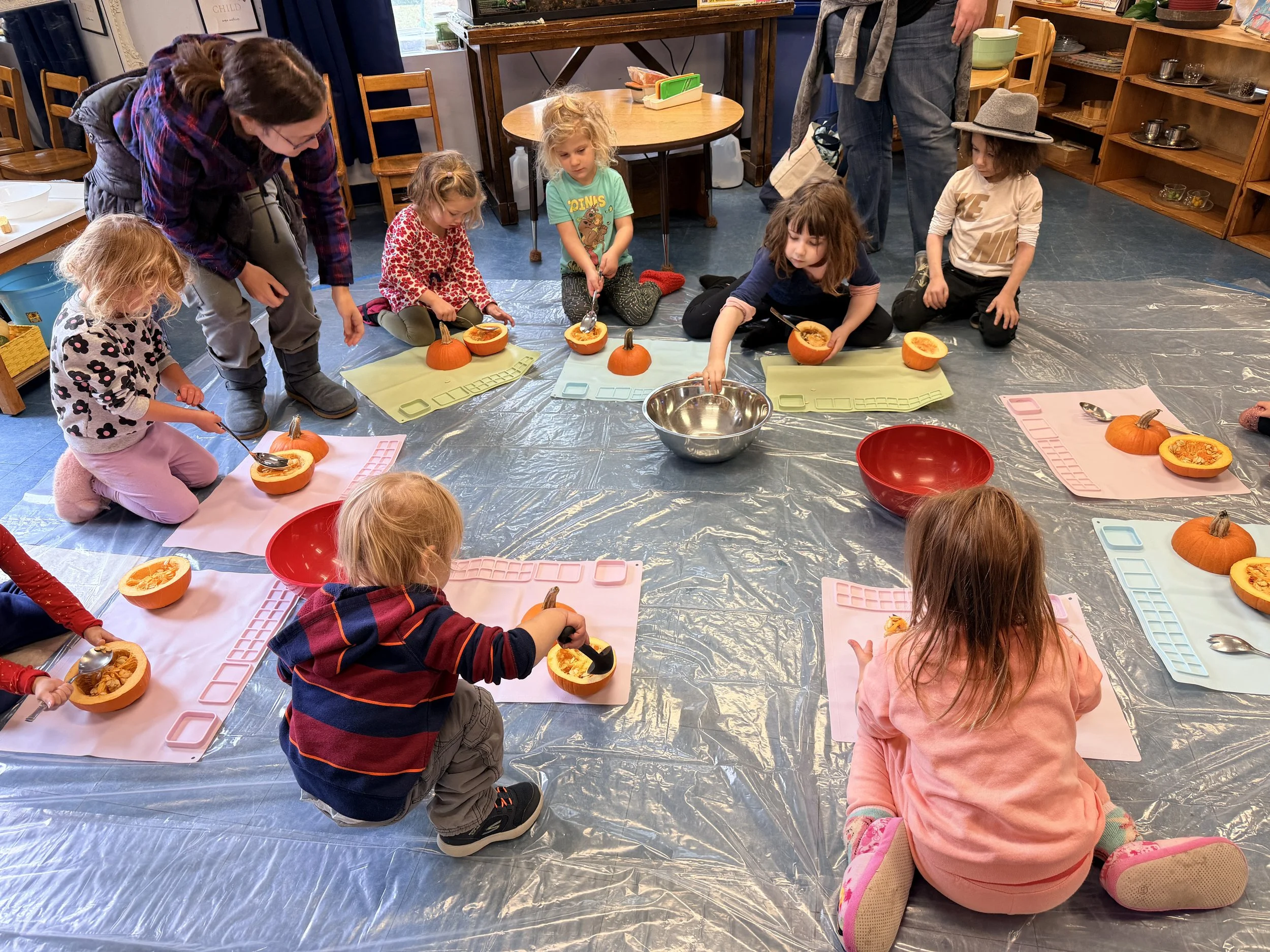 Children sitting on a plastic sheet and carving pumpkins with adult supervision in a room with wooden shelves and tables.