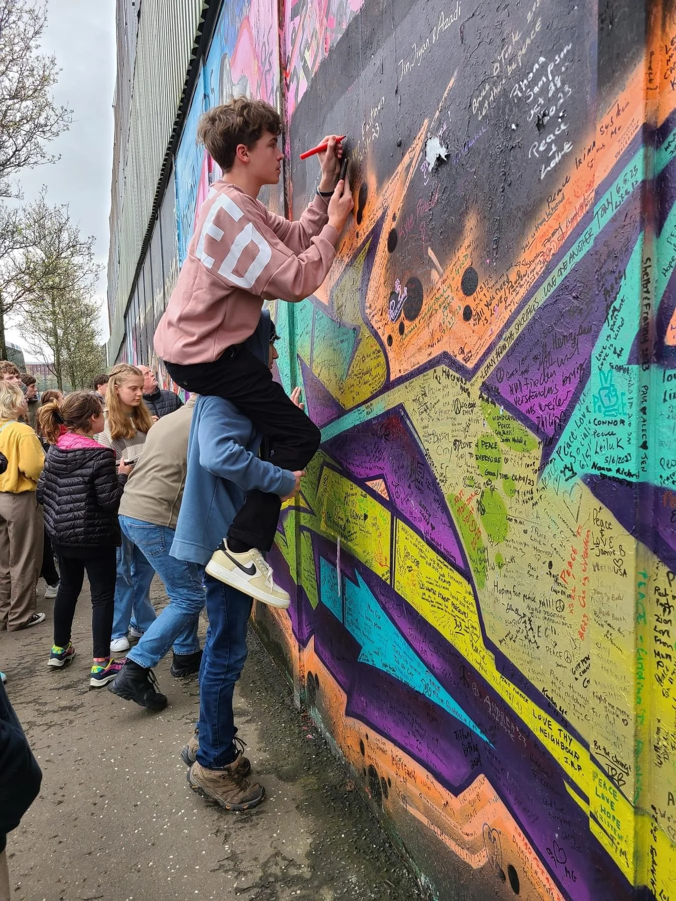 A group of people, including a boy riding on an adult's shoulders, are writing messages on a large, colorful mural covered with writing and drawings in an outdoor setting. The mural features a vibrant, abstract design with multiple colors.