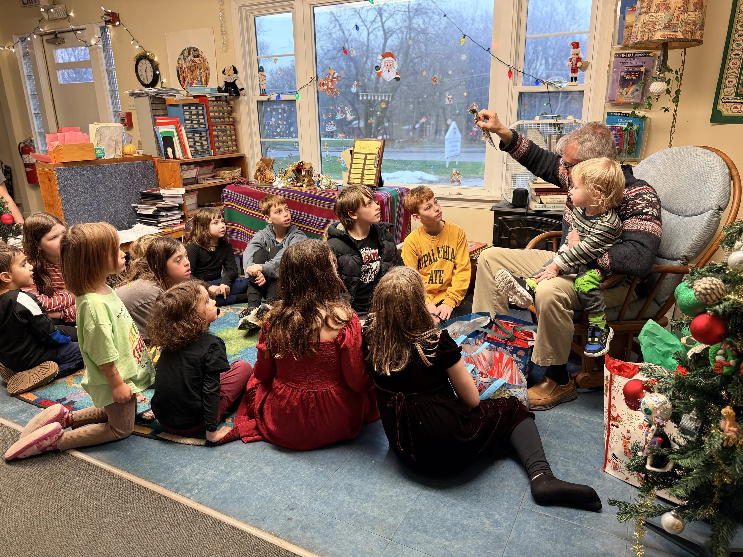 A group of children sitting on a colorful rug, listening attentively to an elderly man who is reading a story to them during a holiday gathering. The room is decorated with Christmas ornaments, fairy lights, and a decorated Christmas tree.
