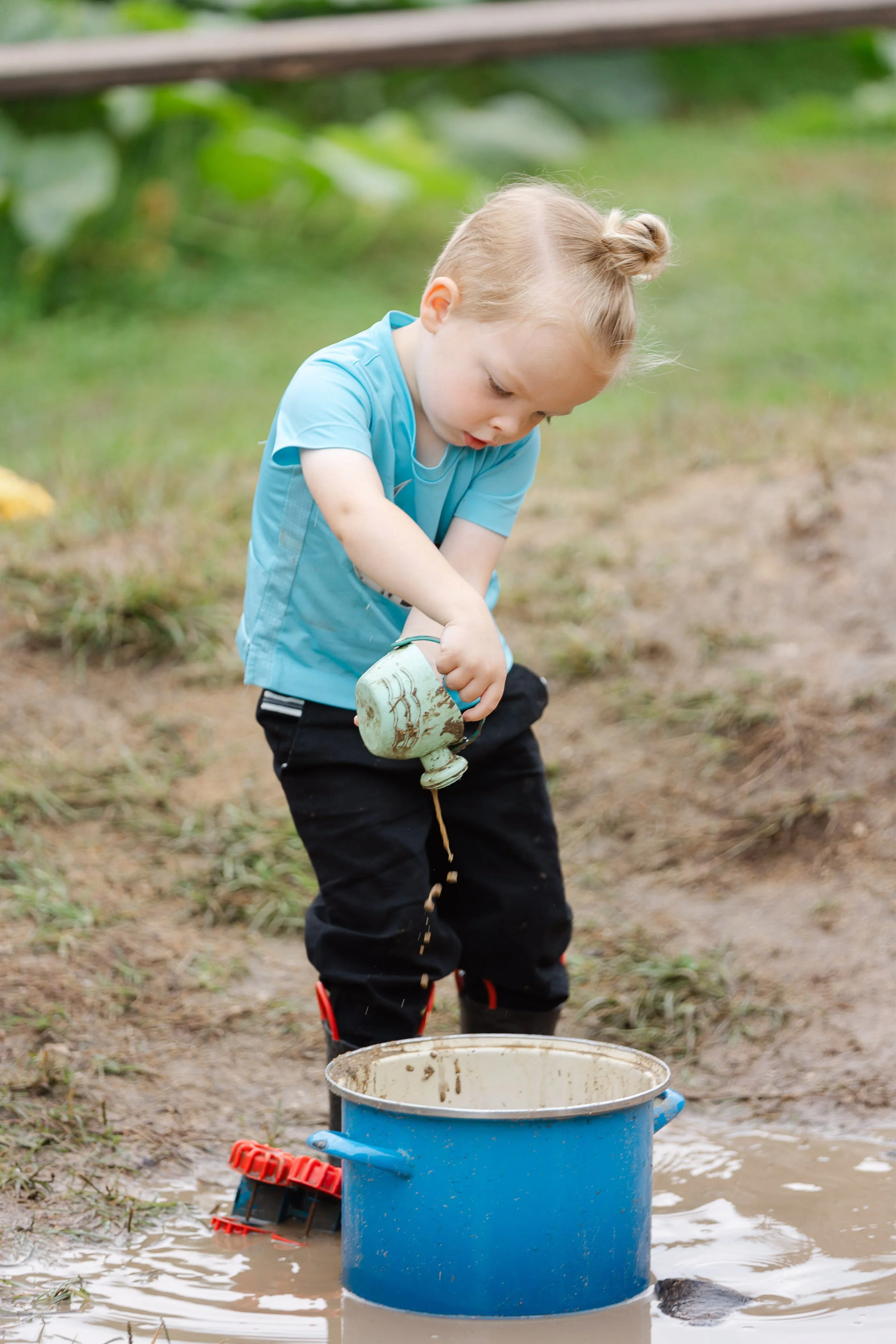 Young girl with blonde hair in a bun, wearing a blue shirt and black pants, playing in a muddy puddle outside, holding a mug with muddy water. There is a large blue bucket in front of her and some toys on the ground.