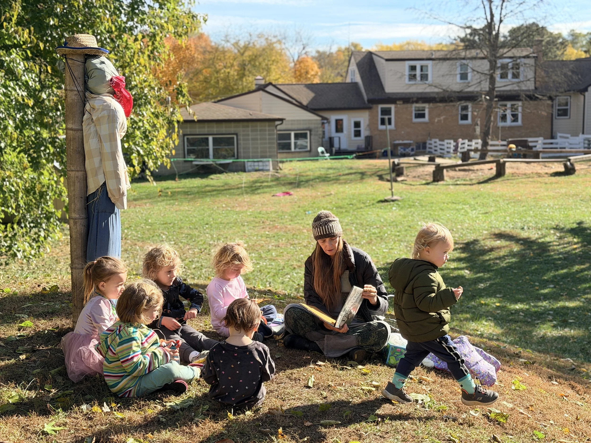 A group of children and an adult sitting on the ground outdoors in a park, with some reading and others looking at objects. A scarecrow dressed in plaid and blue clothing leans against a tree on the left, and houses with fall foliage are in the backg