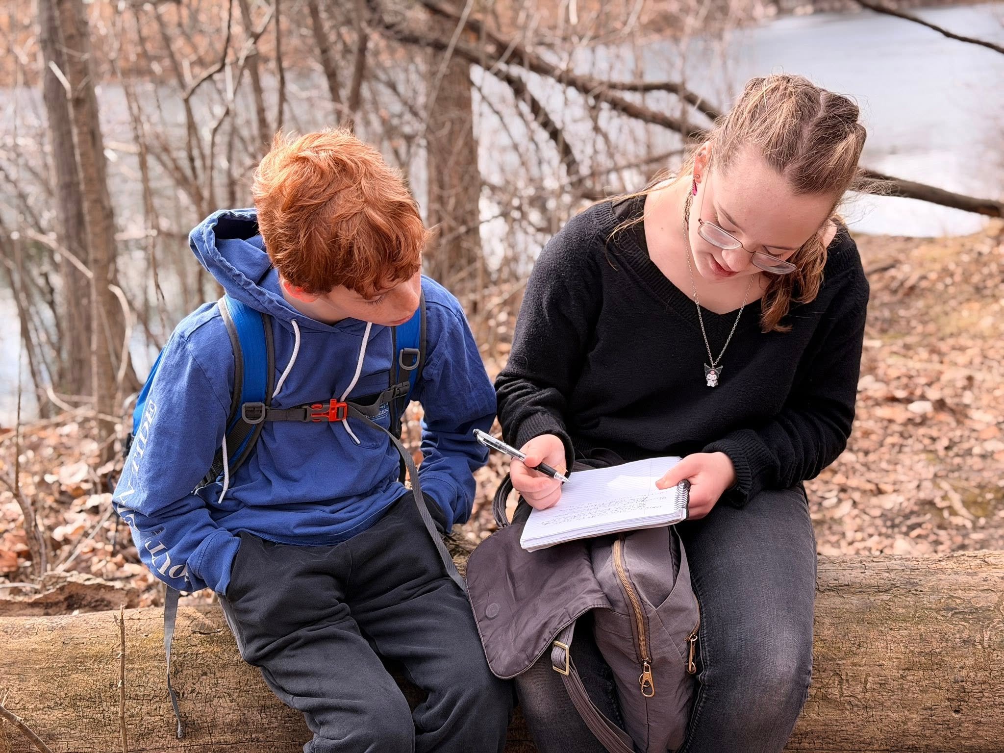 A young woman and a young boy sitting on a log in a forest, looking at a notebook together. The woman is wearing glasses, a black sweater, and has a necklace with a pendant. The boy, who has red hair, is wearing a blue jacket and a backpack.