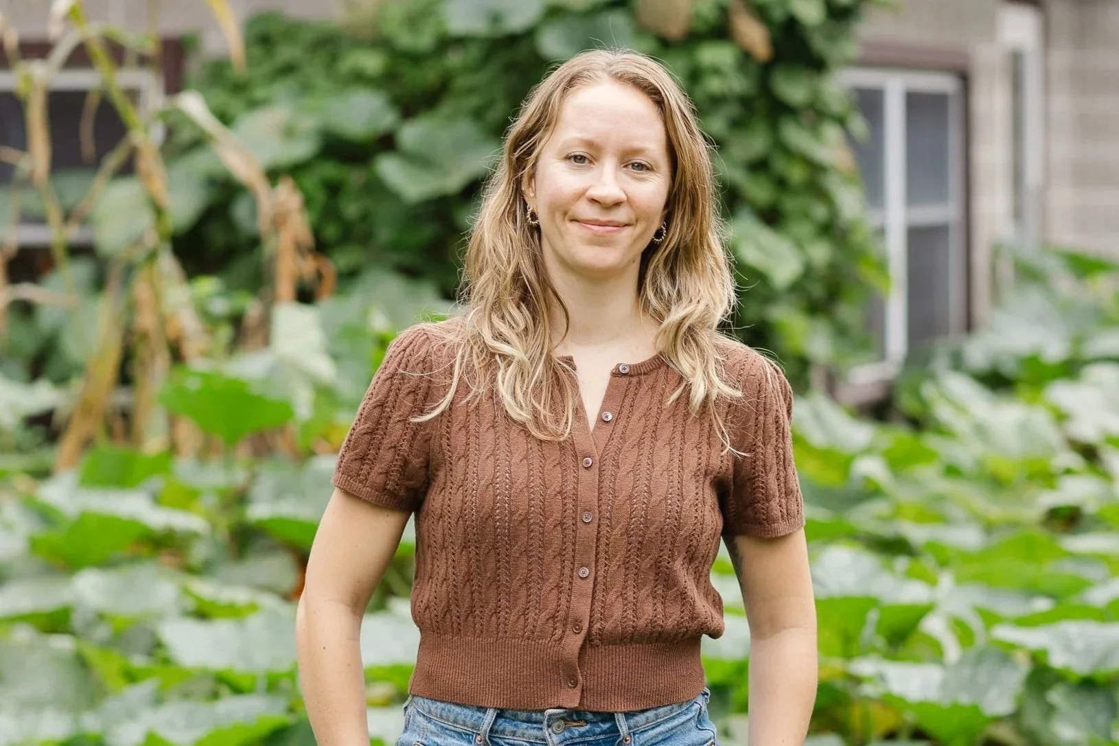 A woman with wavy blonde hair wearing a brown short-sleeve knit shirt and blue jeans, standing outdoors in front of green leafy plants and a building with windows.