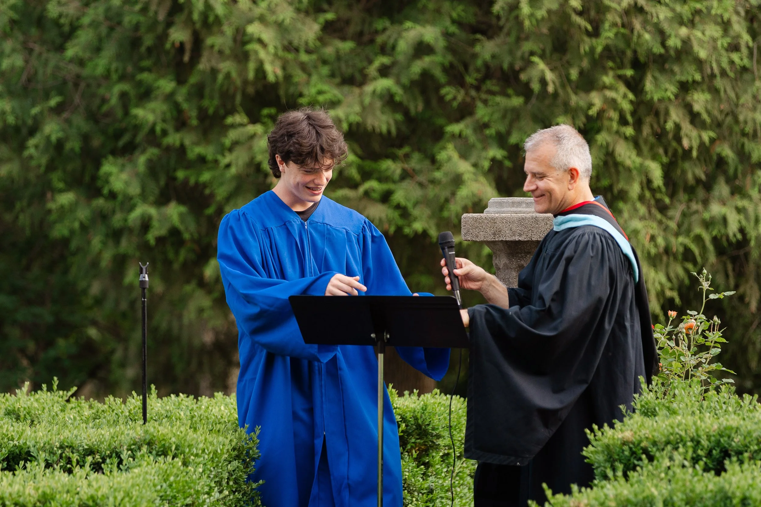 A young man in a blue graduation gown and an older man in academic robes are smiling during a graduation ceremony outdoors. The young man is receiving a diploma from the older man, who is holding a microphone and standing on a grassy area with lush g